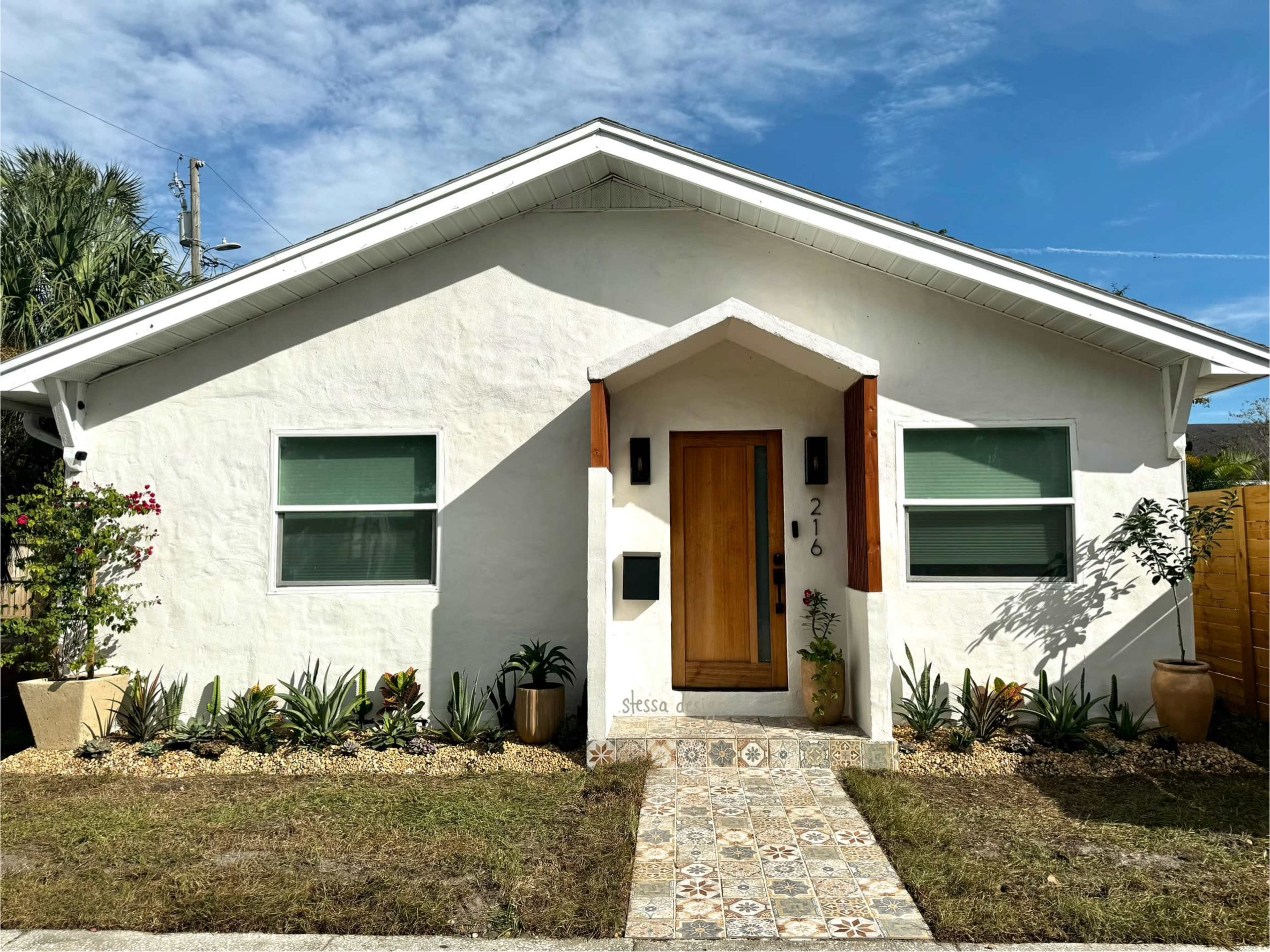 A white stucco house with a wooden front door and landscaped yard, featuring a decorative pathway and garden plants.