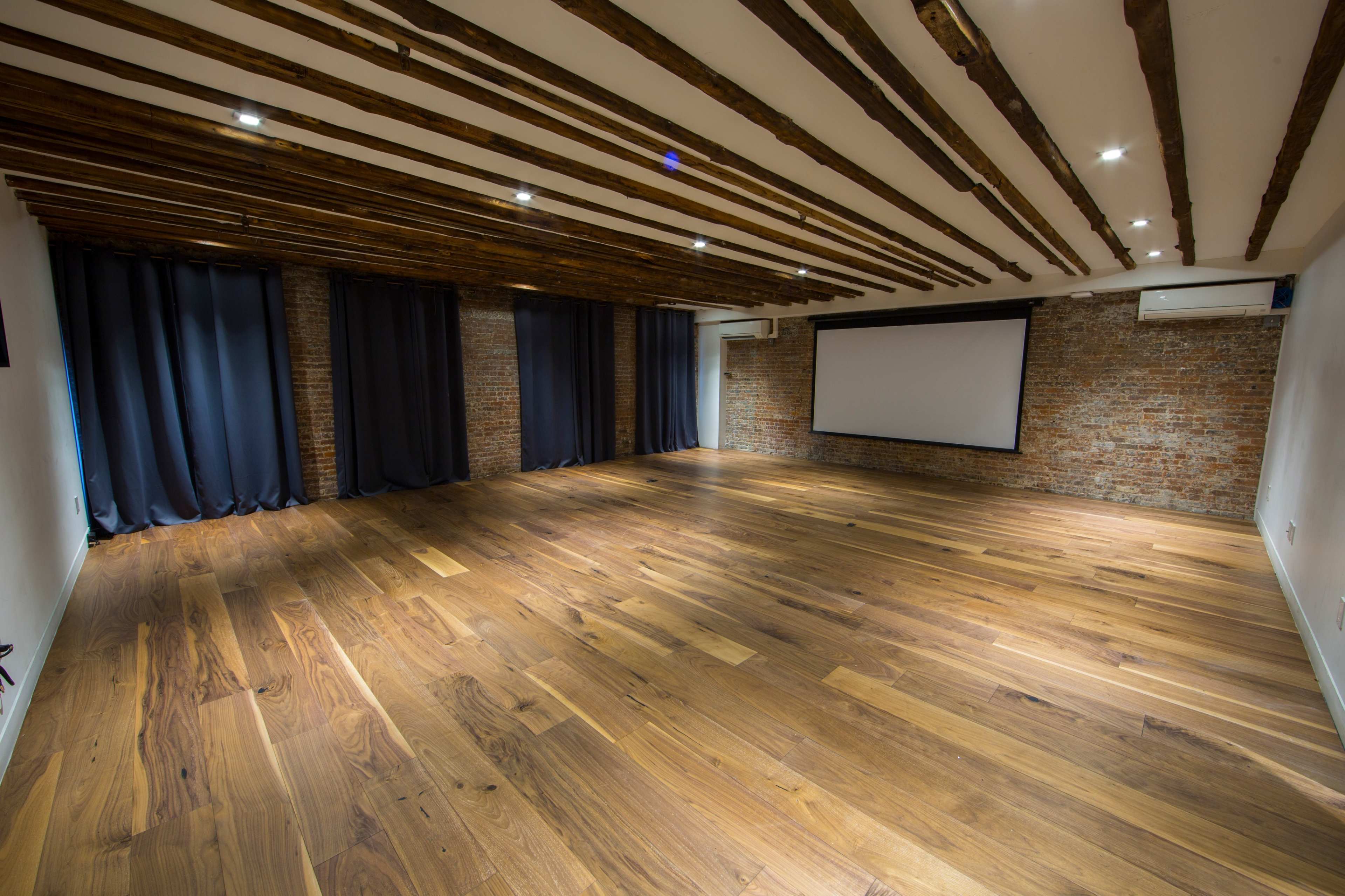 A spacious interior room with wooden flooring, exposed brick walls, and beams on the ceiling, featuring dark curtains and a projection screen.