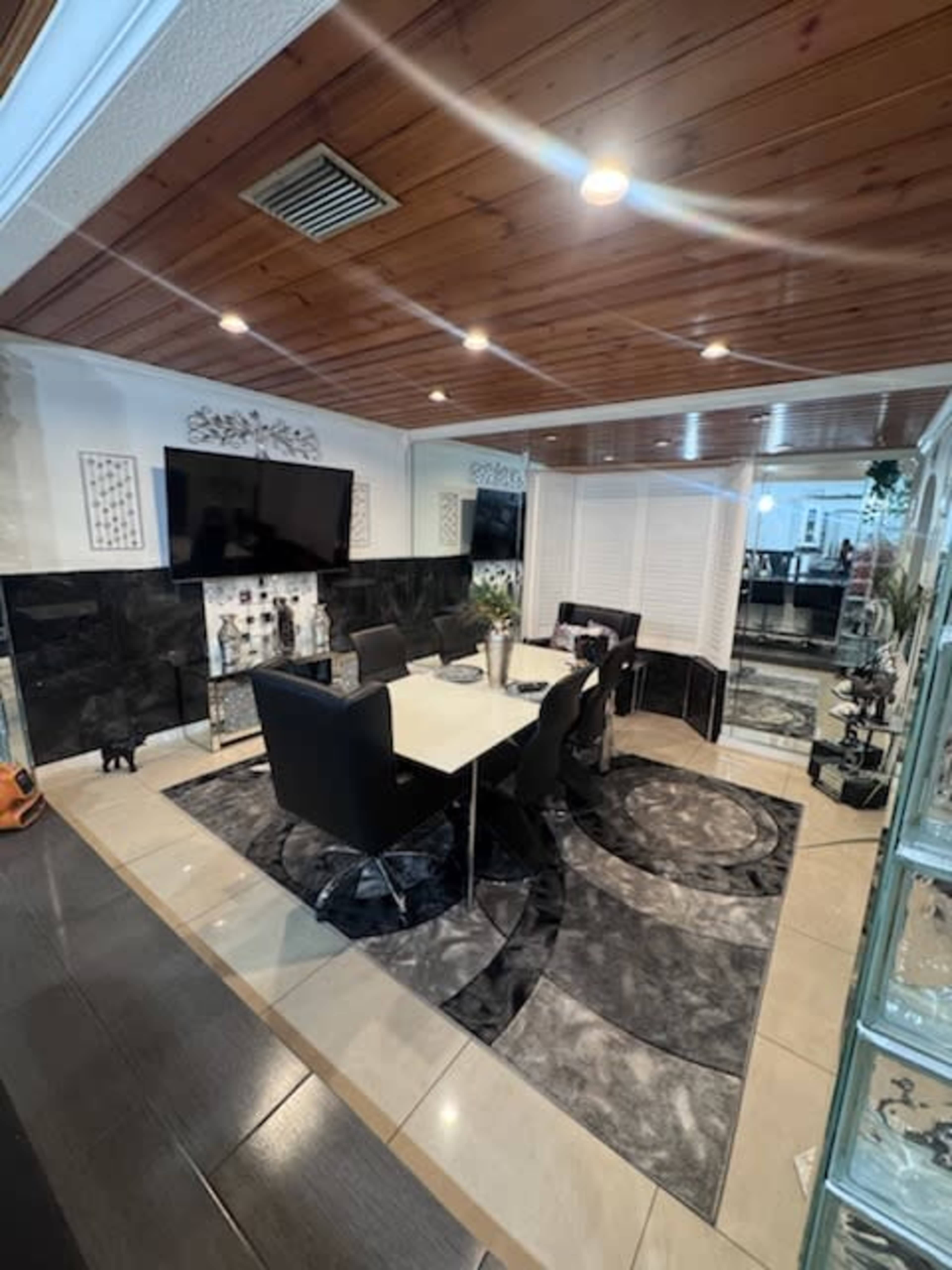 The image shows a modern dining area with a table surrounded by black chairs, a patterned rug, and a large flat-screen TV mounted on the wall.