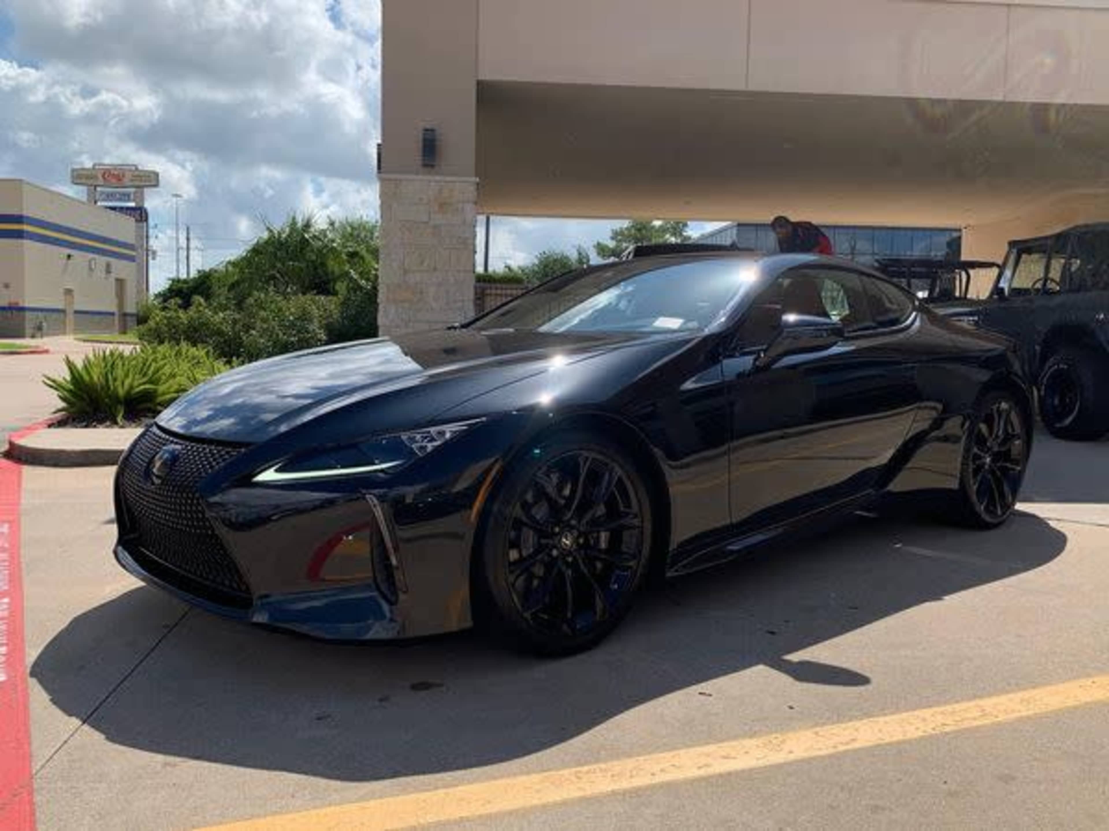 A black sports car is parked outside a building on a paved surface under a partly cloudy sky.