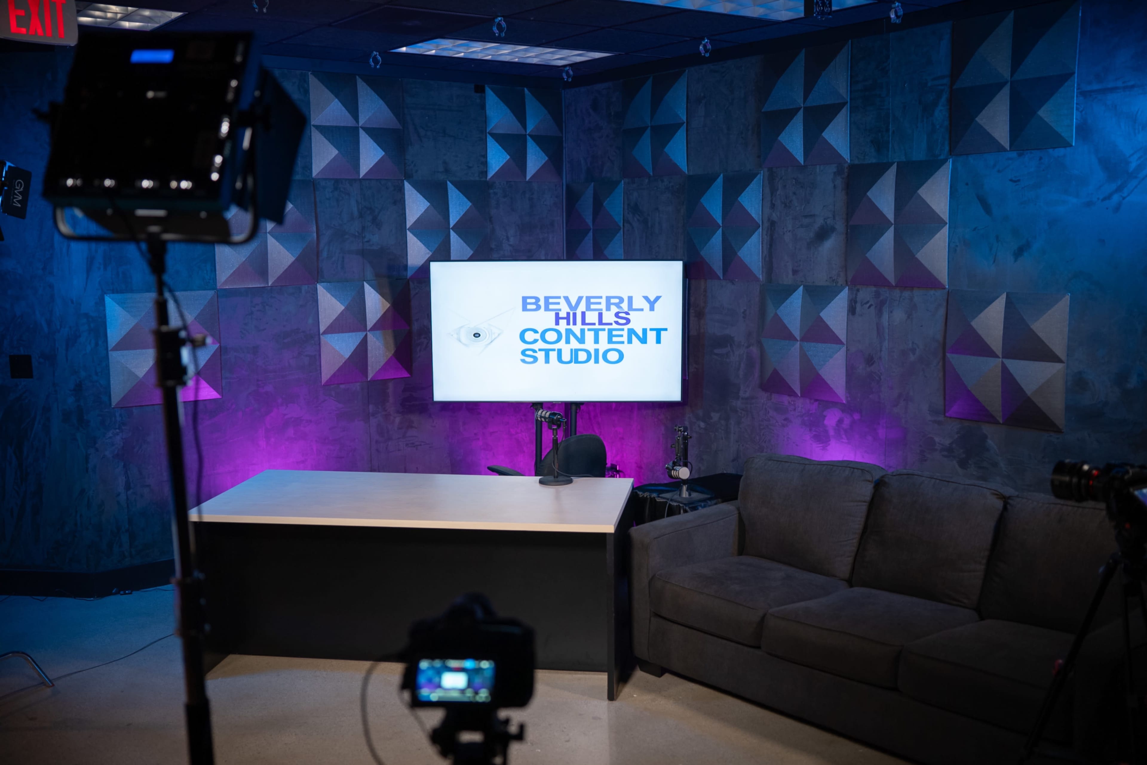The image shows a well-lit studio setup featuring a large screen displaying the text "BEVERLY HILLS CONTENT STUDIO," a desk, and a gray sofa against a textured wall with geometric patterns.