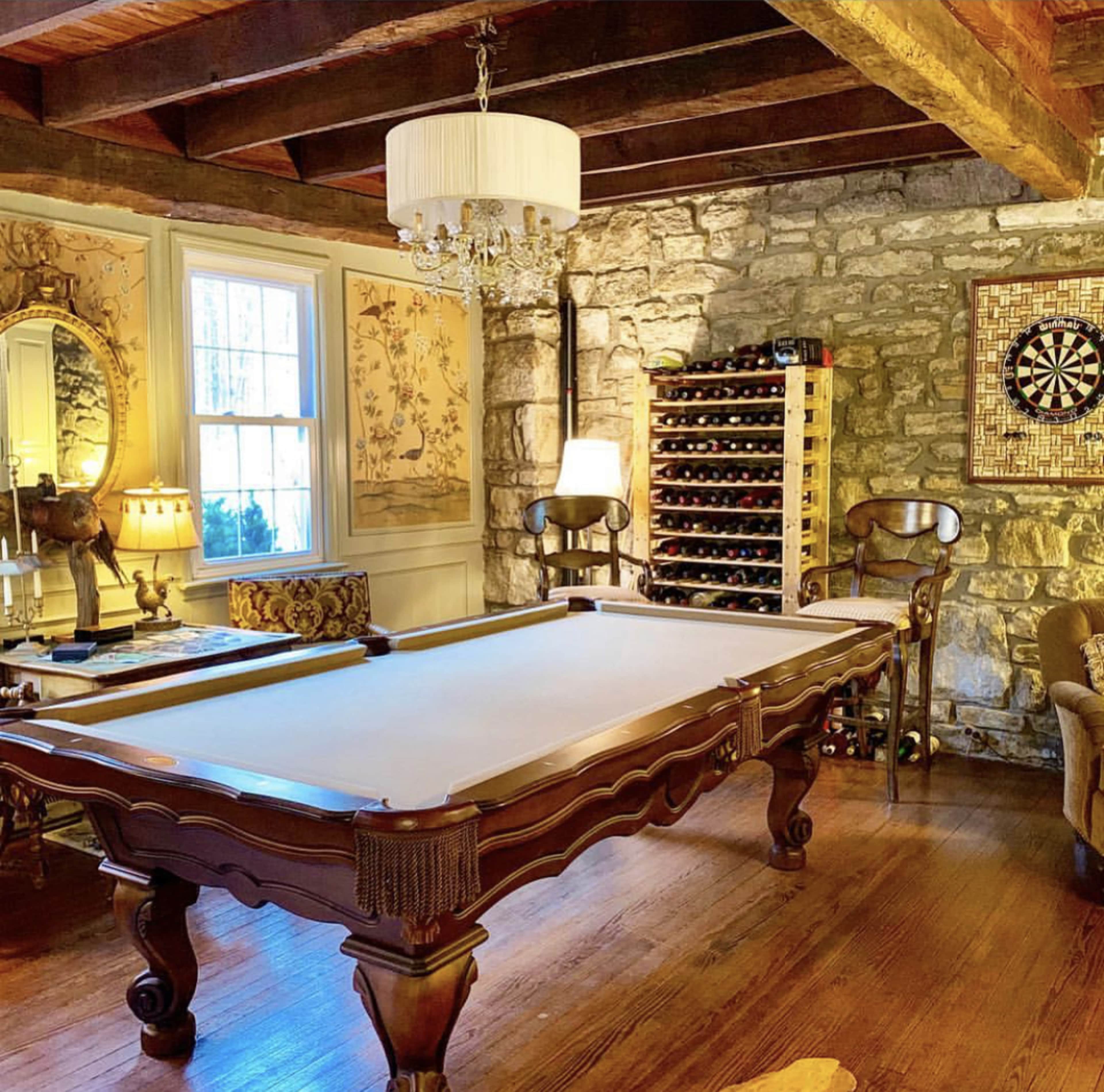 The image shows a game room featuring a billiards table, a wine rack against a stone wall, and decorative furnishings.