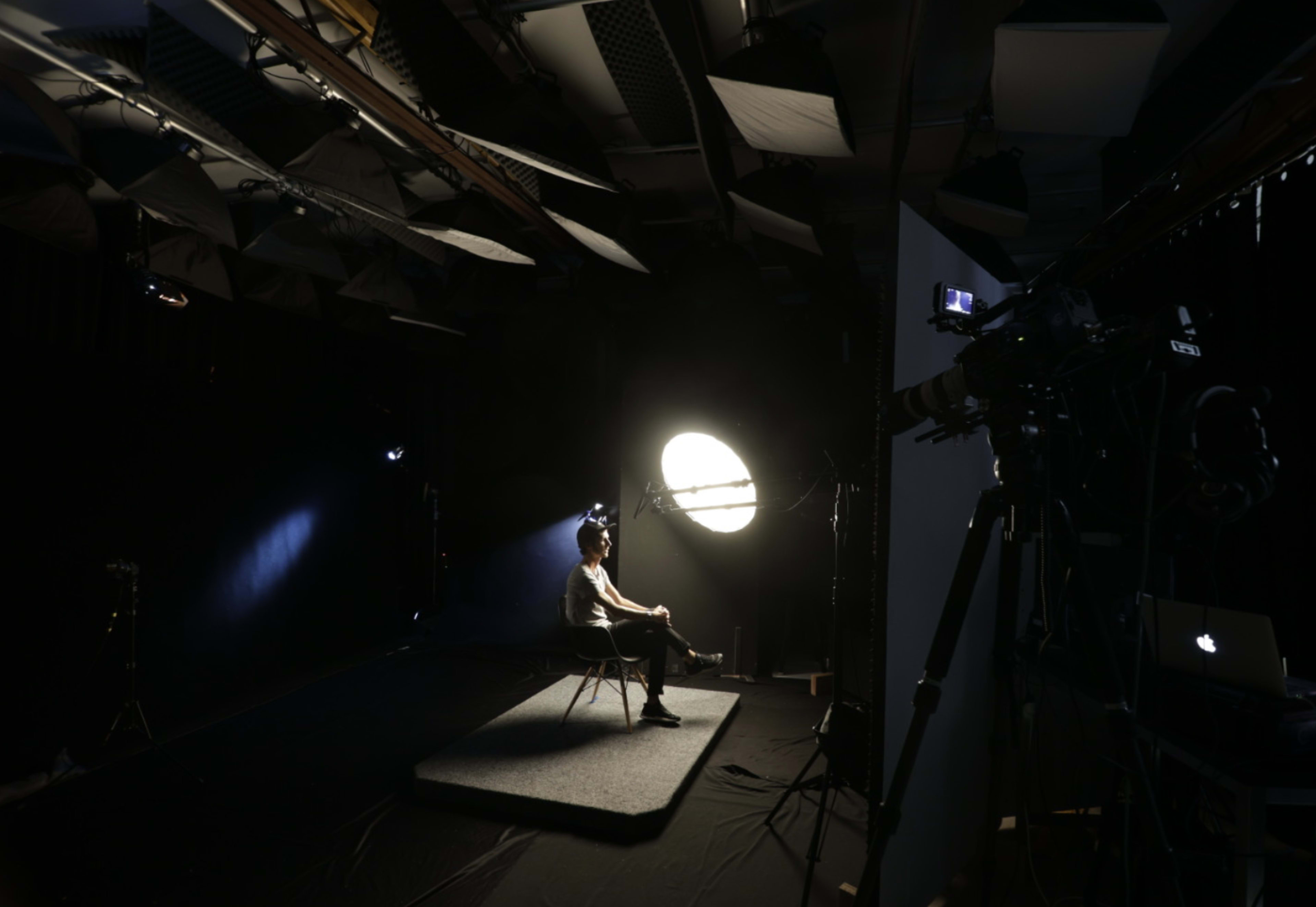 A person sits on a chair in a dimly lit studio with a large circular light illuminating them, surrounded by cameras and lighting equipment.