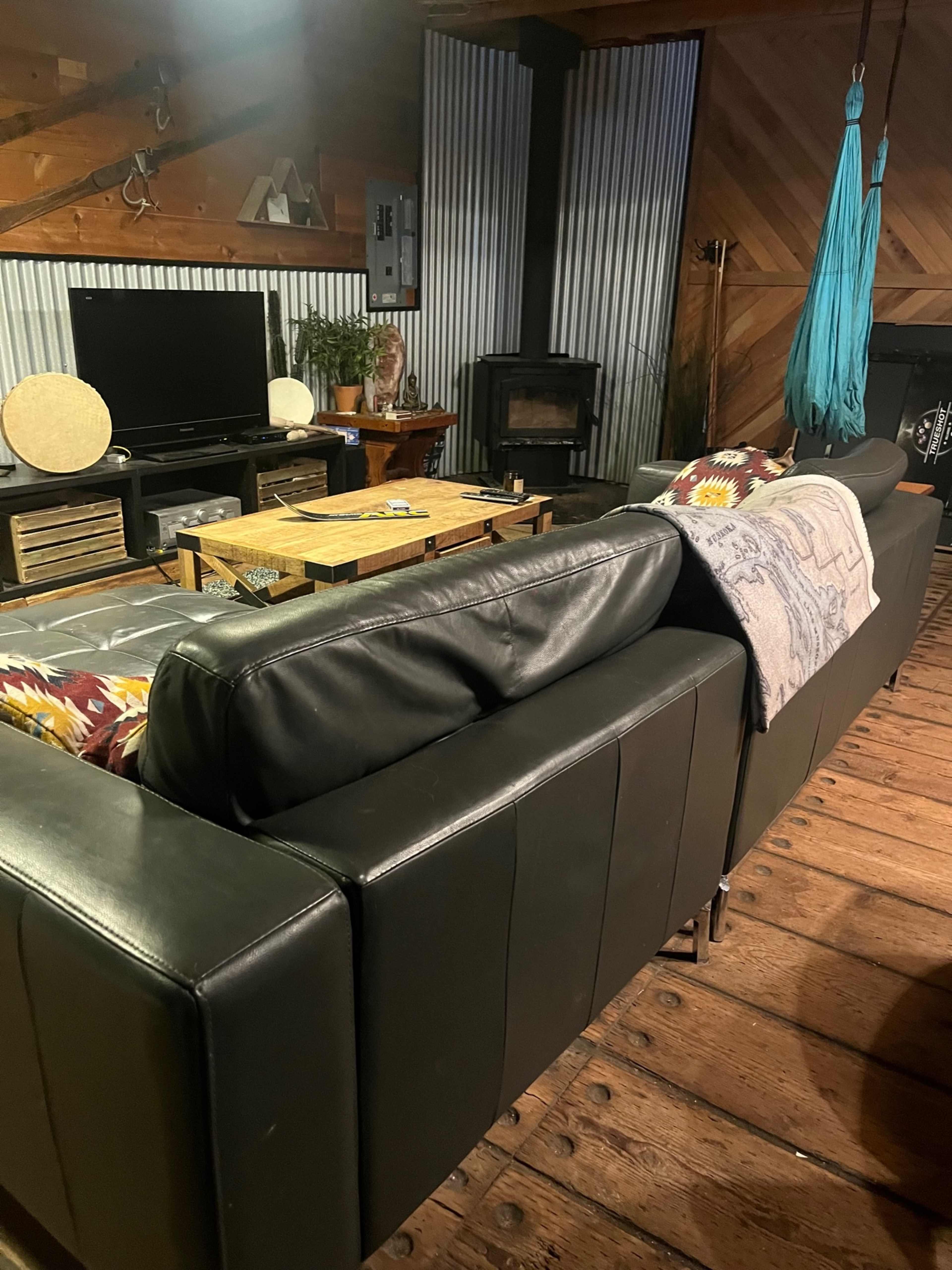 A living room with a black leather sofa facing a wooden coffee table, a television unit, and a wood stove against a backdrop of corrugated metal and wooden walls.