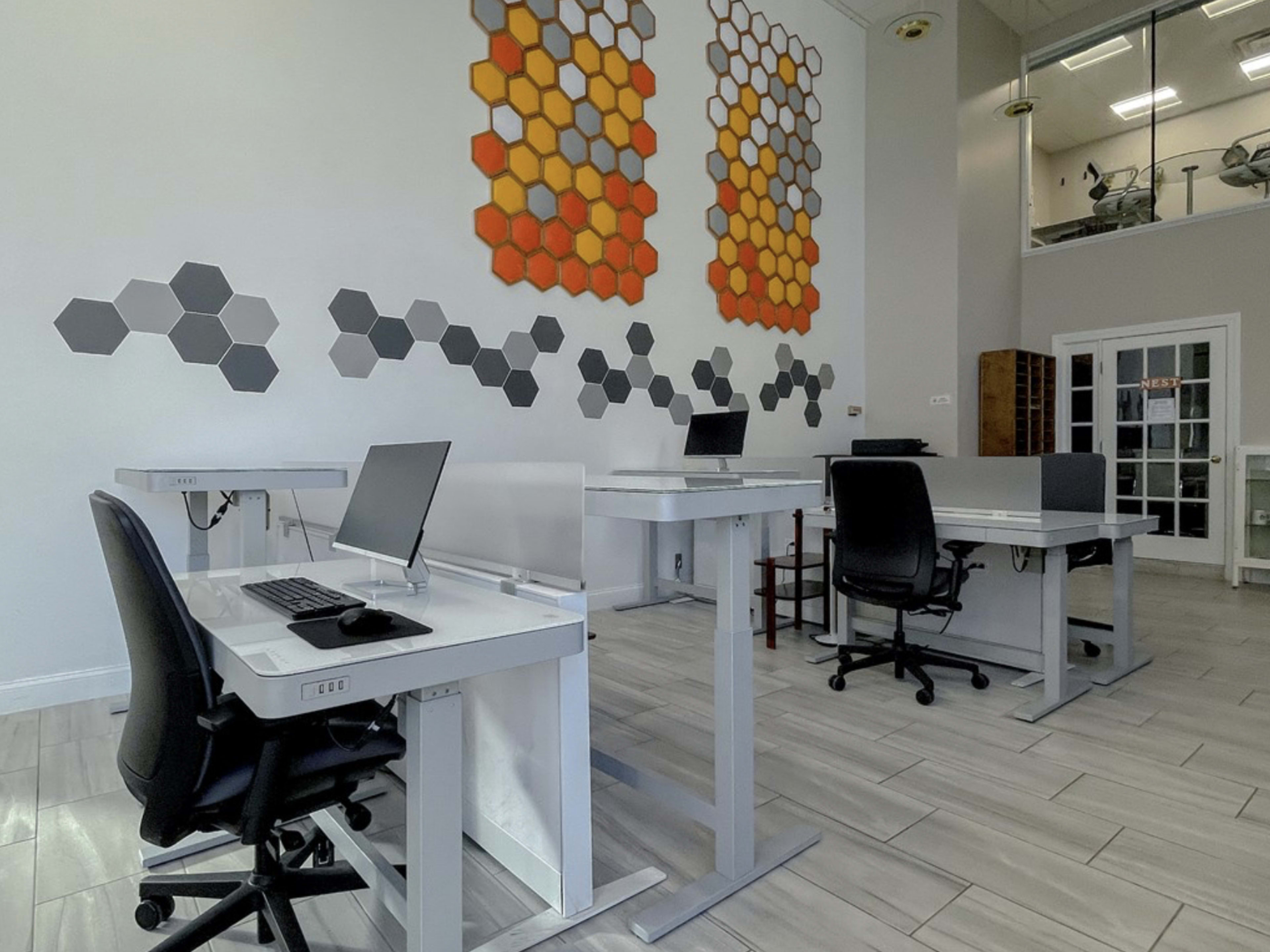 A modern office space with several workstations featuring adjustable desks and computers, accompanied by a wall decoration of hexagonal patterns in orange and gray.