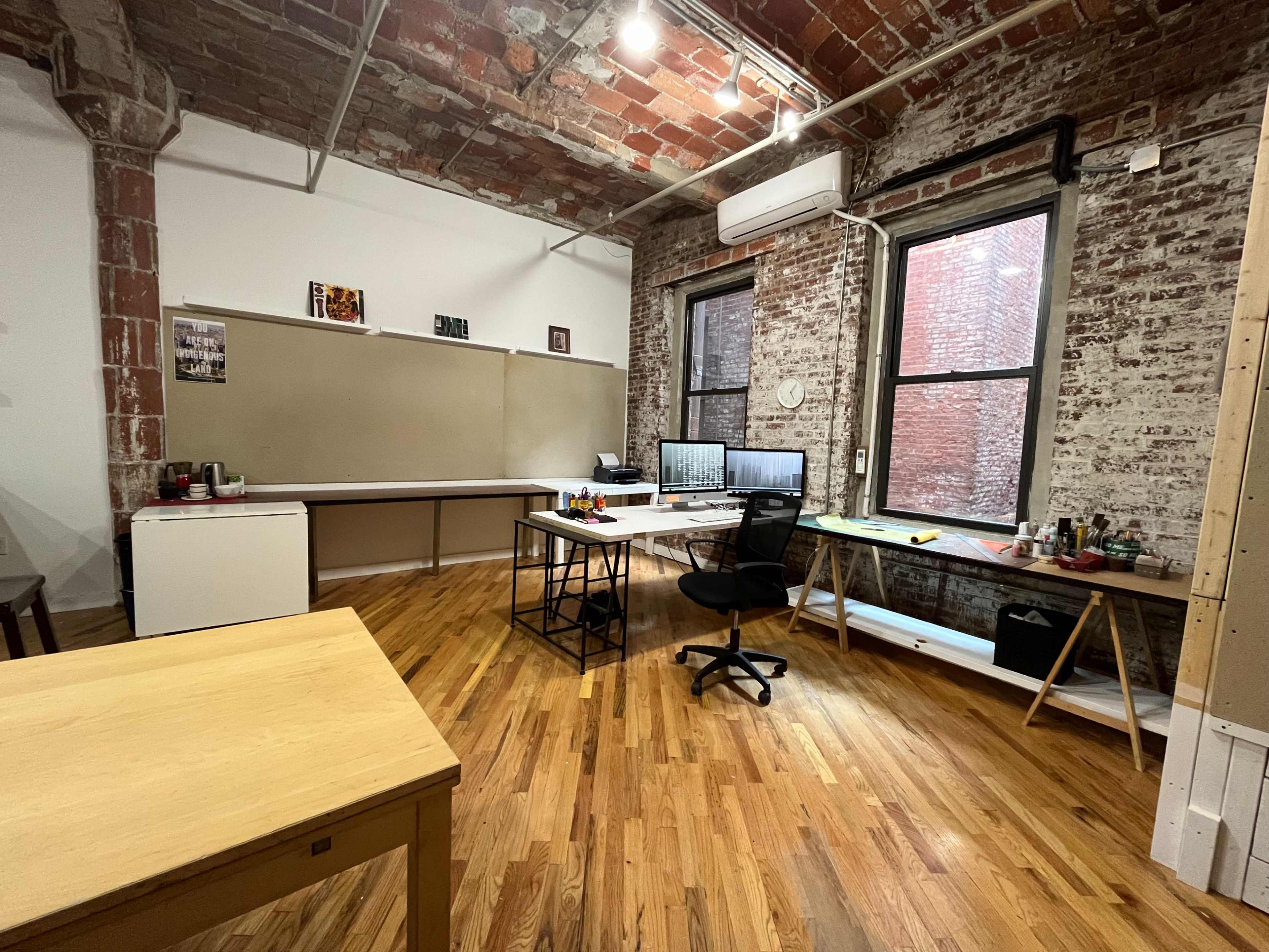 A spacious office with exposed brick walls, wooden floors, and modern furniture, including a desk and a chair arranged near large windows.