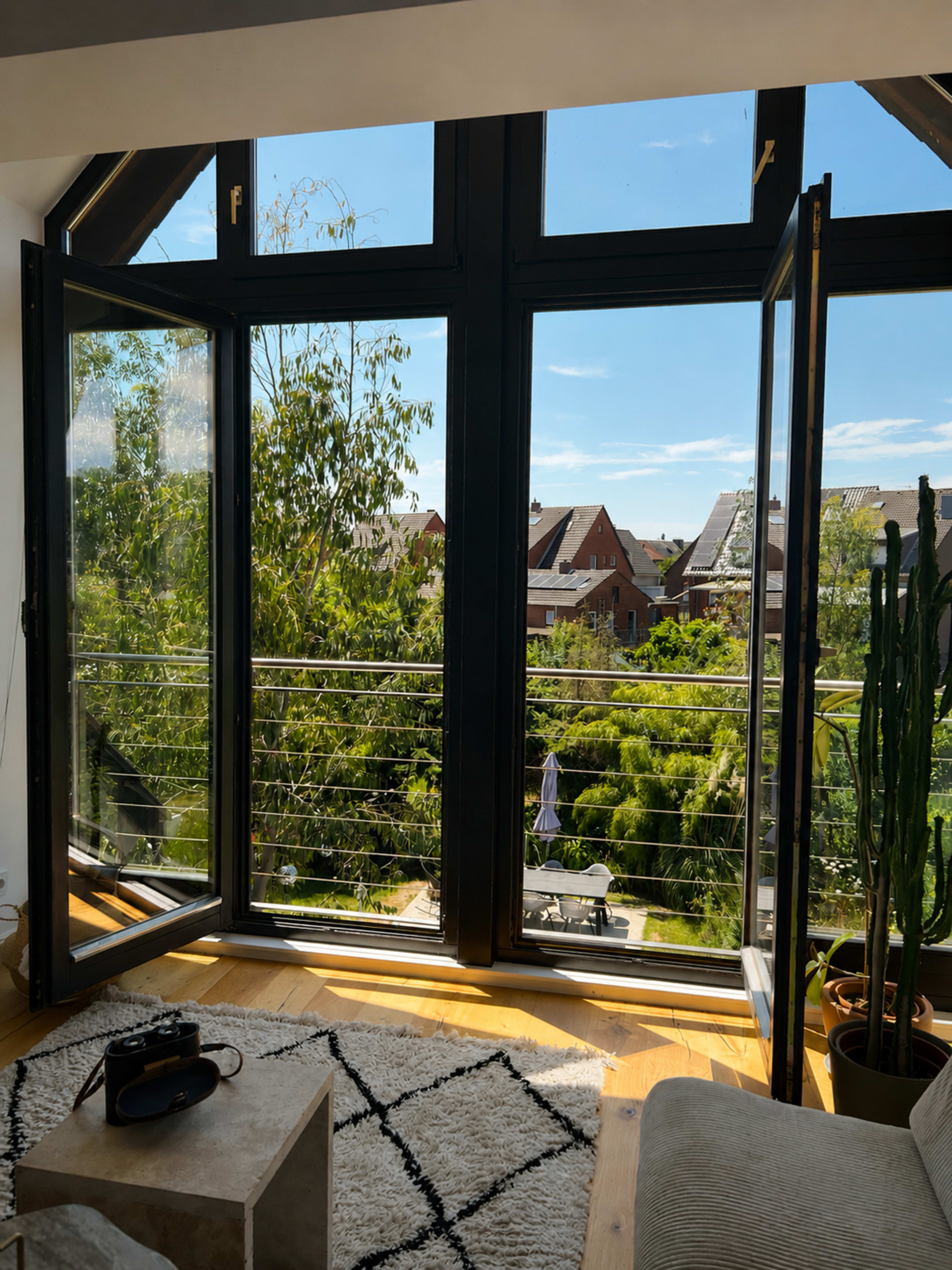 The image shows a modern living space with large open windows overlooking a green area and nearby rooftops.