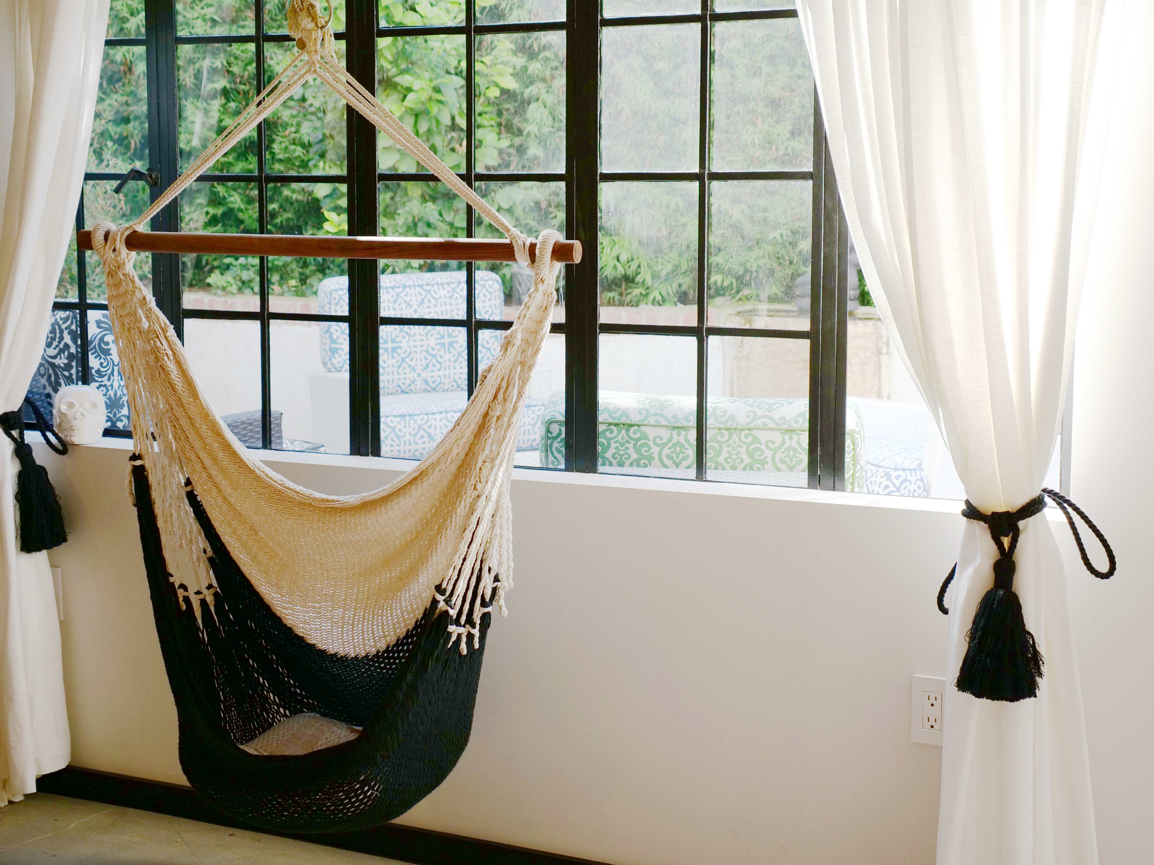 A hammock hangs from a wooden bar in a room with large windows, revealing a view of an outdoor area with patterned furniture.