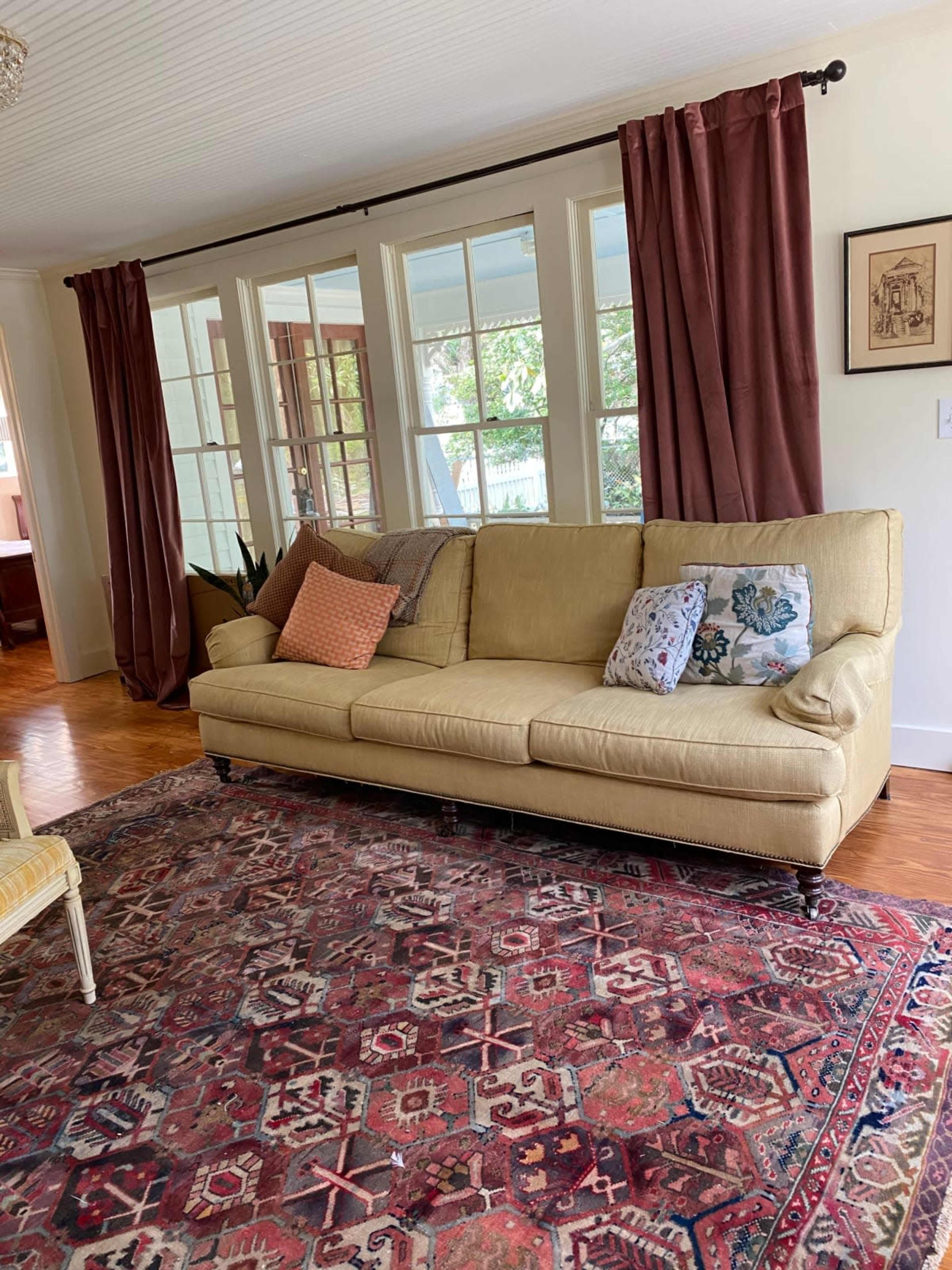 A light-colored sofa with decorative pillows is positioned on a patterned rug in a room with large windows and draped curtains.