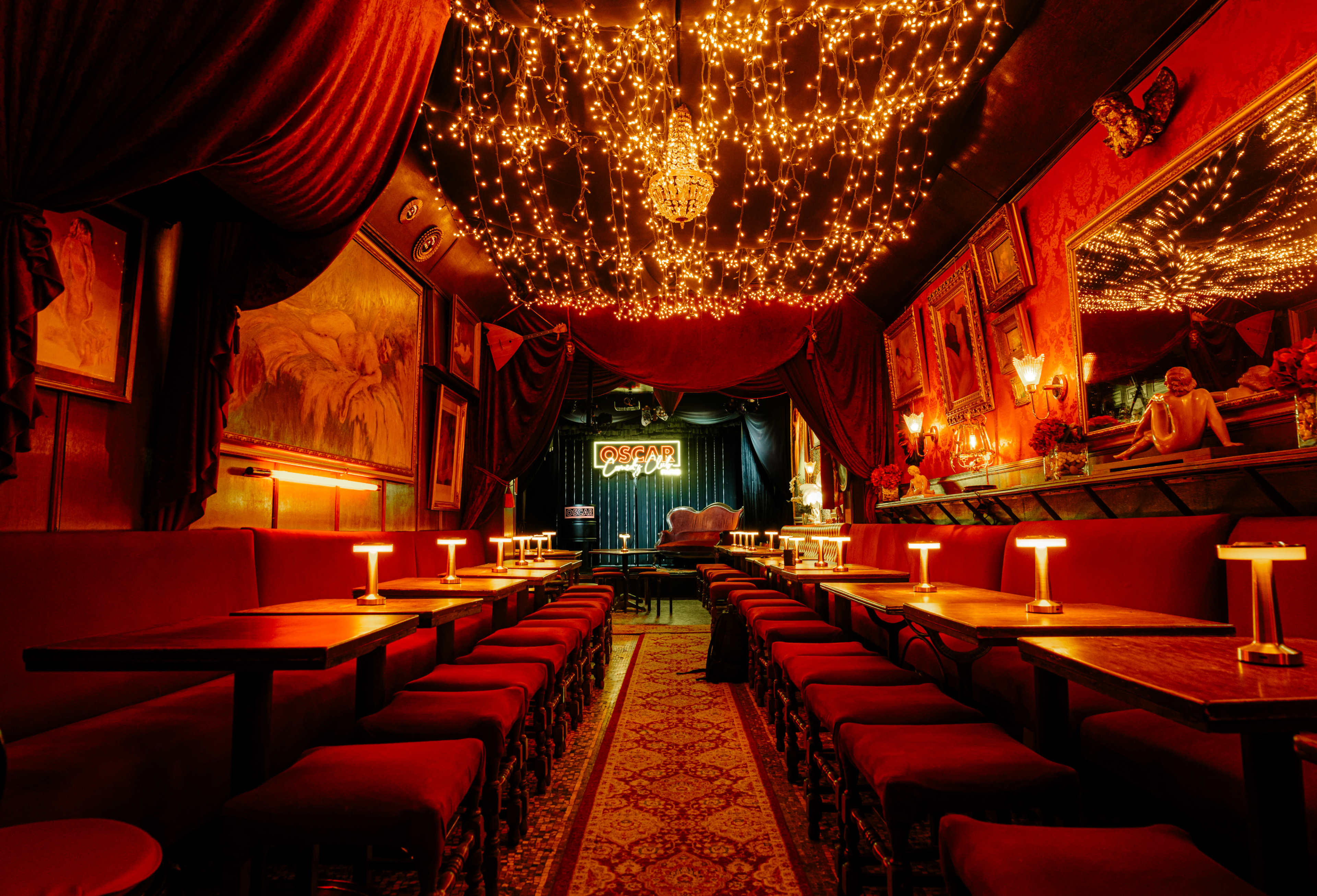 The image shows a warmly lit lounge with a long row of red upholstered tables and chairs, embellished with hanging string lights and rich décor on the walls.