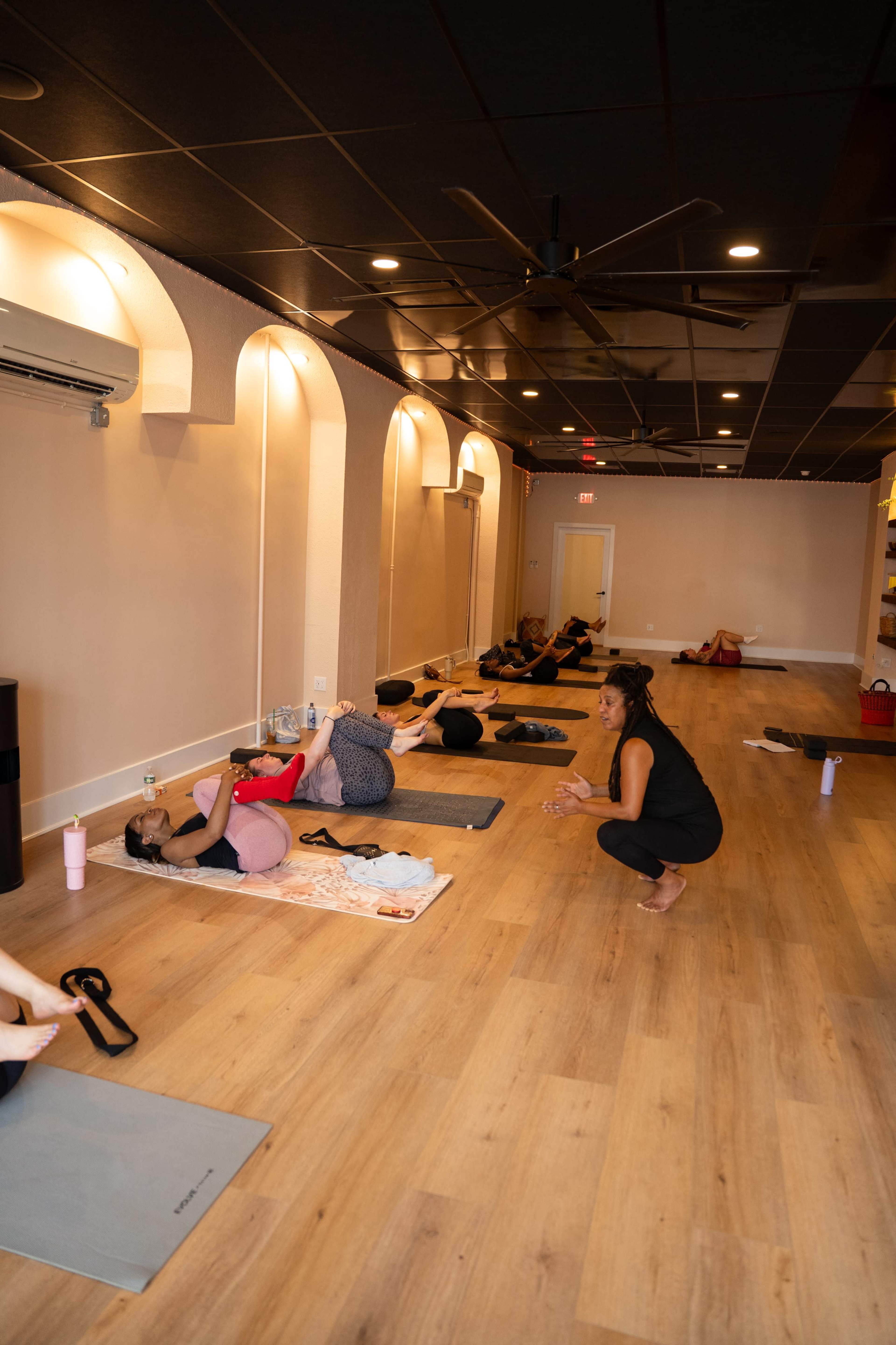 The image shows a yoga studio with several individuals practicing yoga on mats while a teacher observes and instructs.