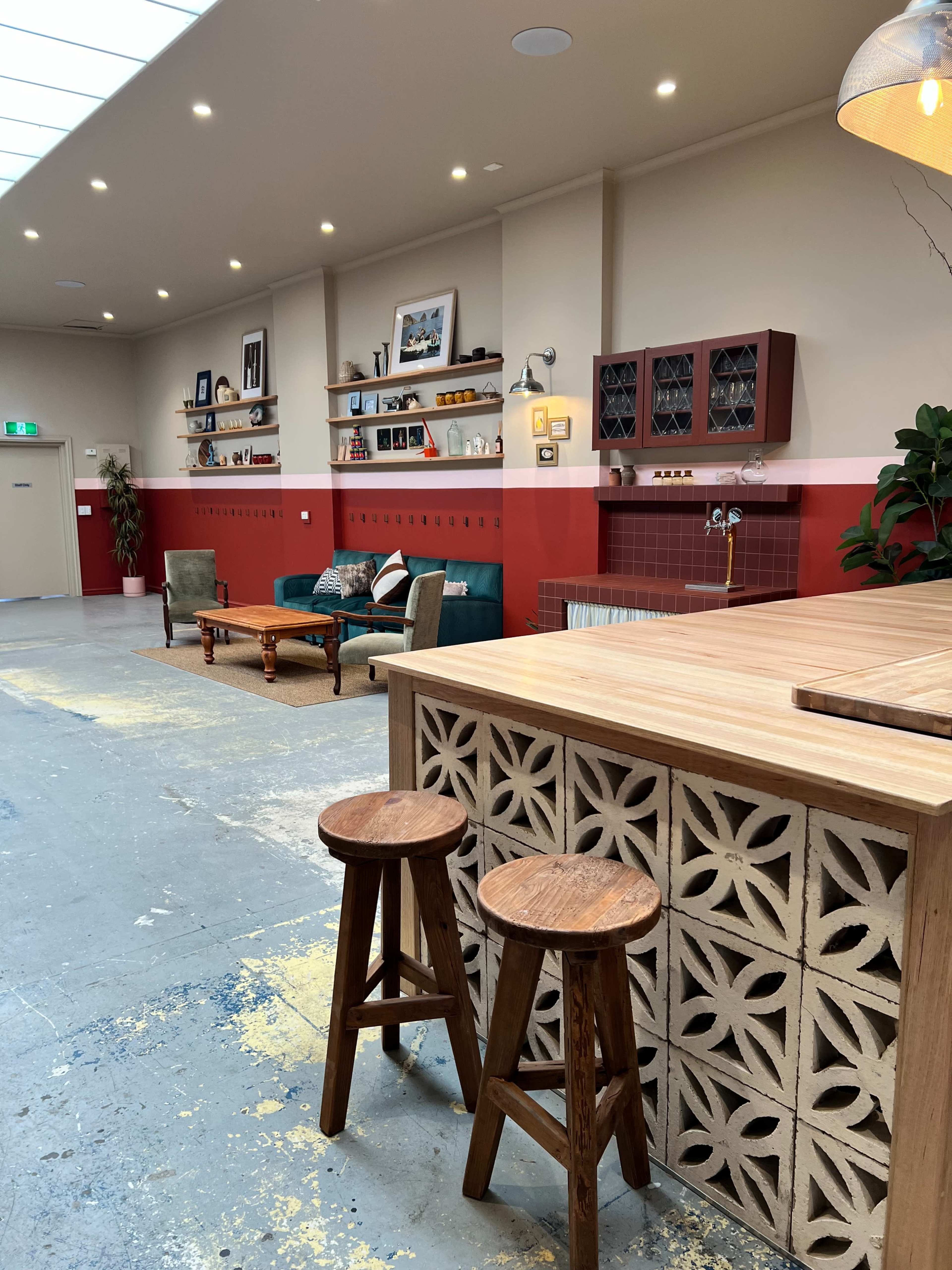 The image shows a spacious interior with a wooden bar featuring decorative panels, two wooden stools, a green sofa, and various shelves adorned with pictures and decorative items.