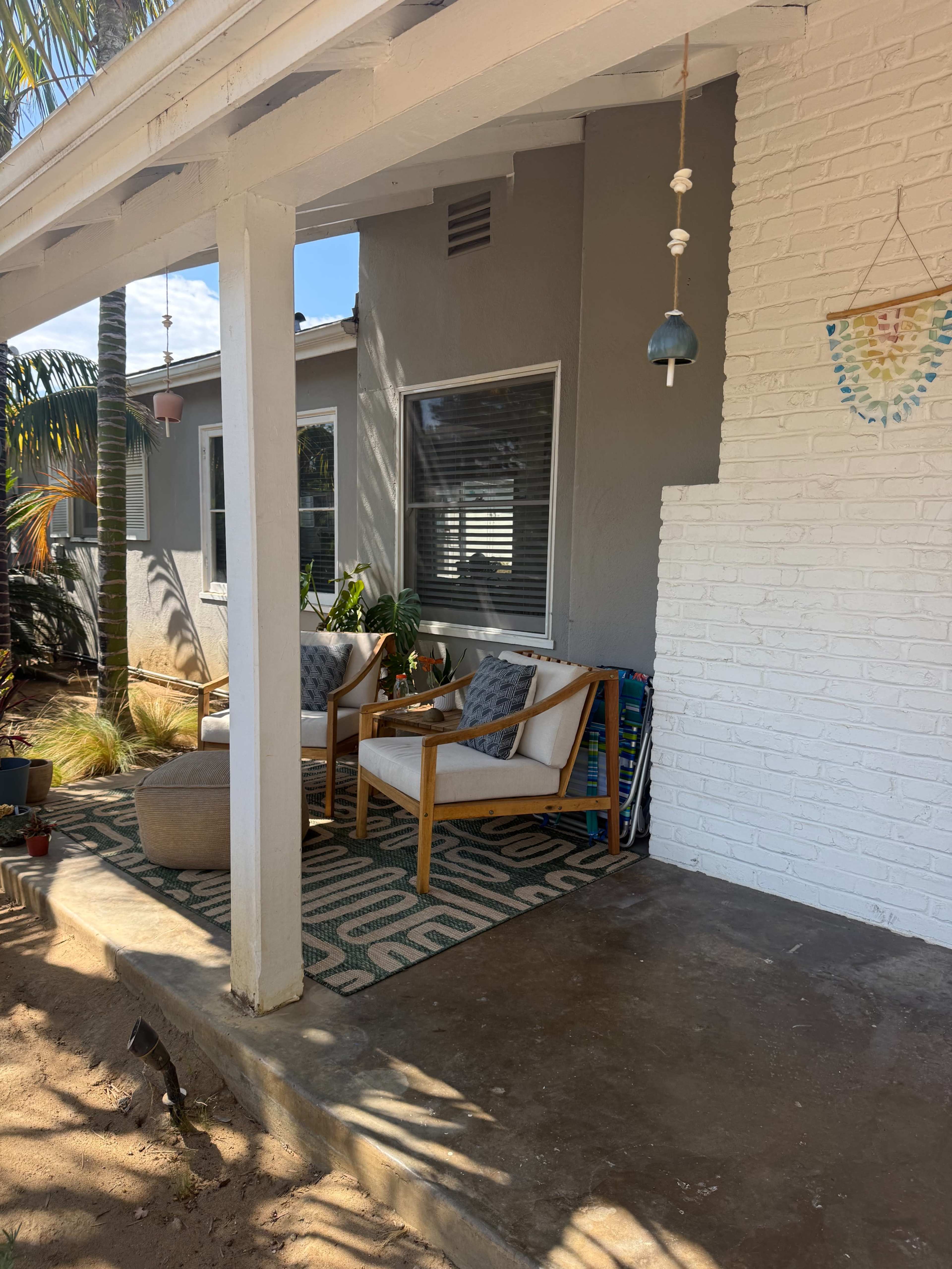 The image depicts a cozy porch with two armchairs, a round ottoman, and a patterned rug under a white overhang.