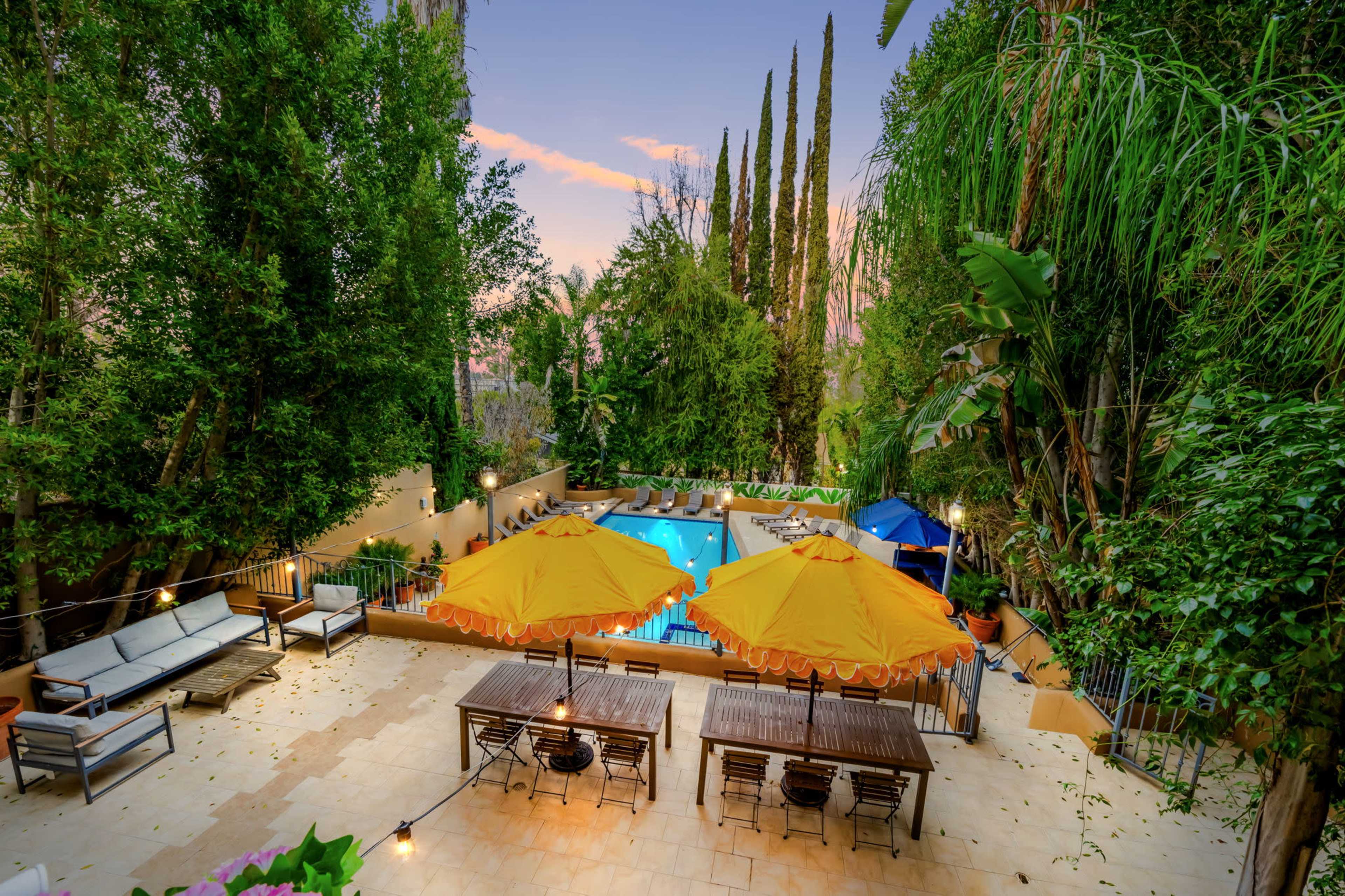 A backyard garden with a swimming pool, surrounded by tall trees, outdoor furniture, and two large orange umbrellas over a dining area.