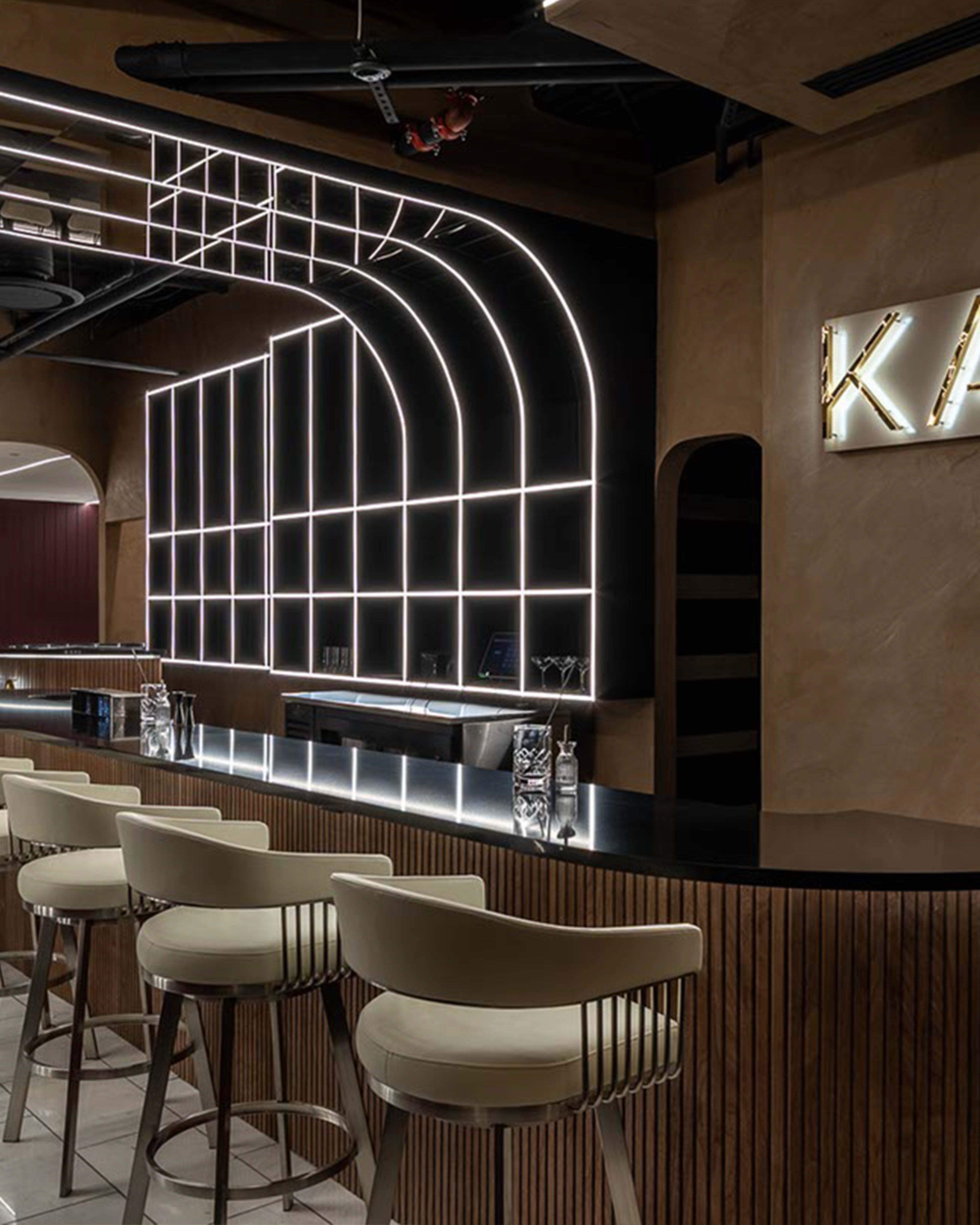 The image shows a modern bar interior with a sleek counter, padded stools, and a back wall featuring intricate geometric LED lighting.