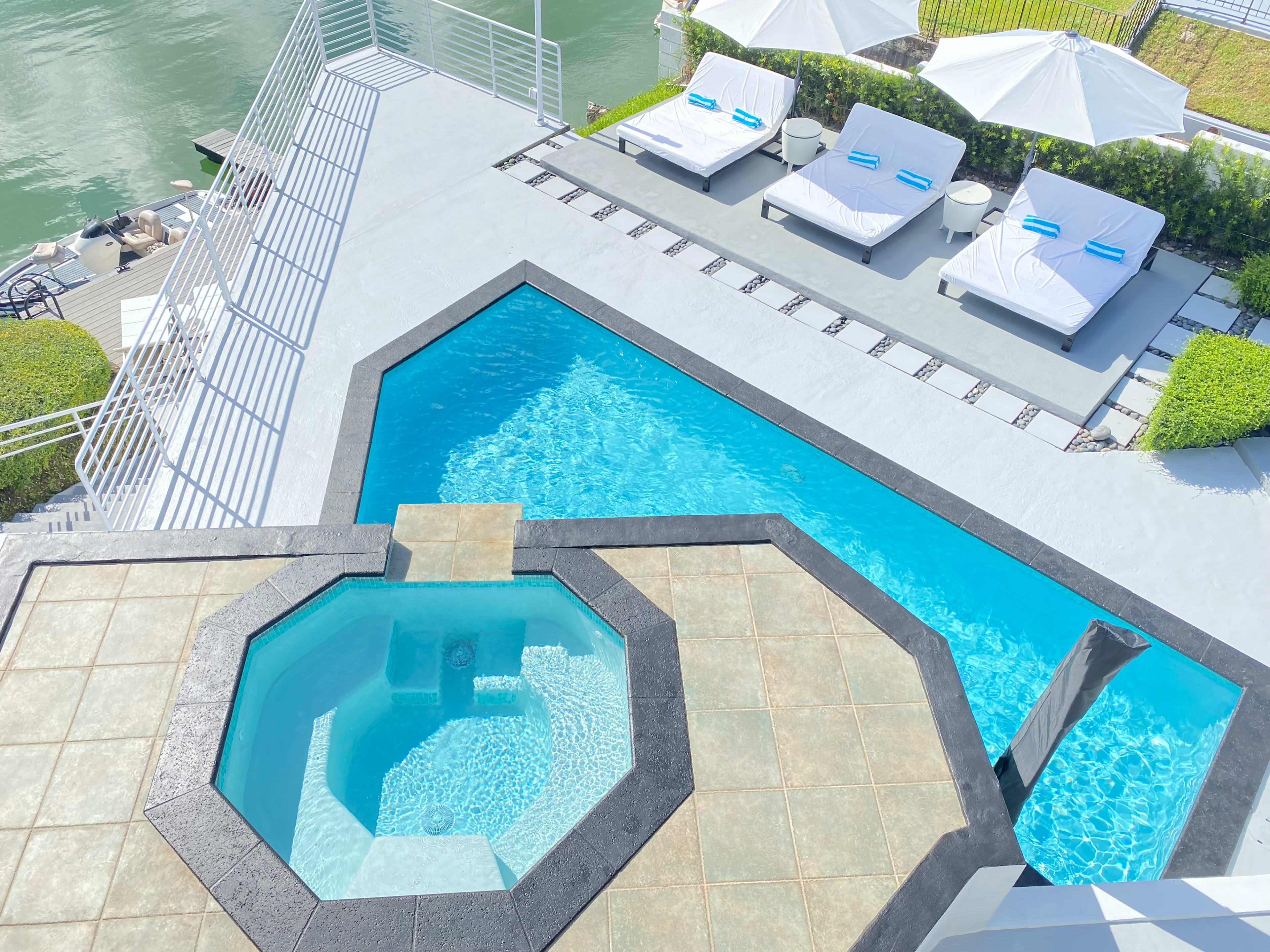 The image shows a modern pool area featuring a large rectangular swimming pool, a hot tub, and several lounge chairs under white umbrellas.