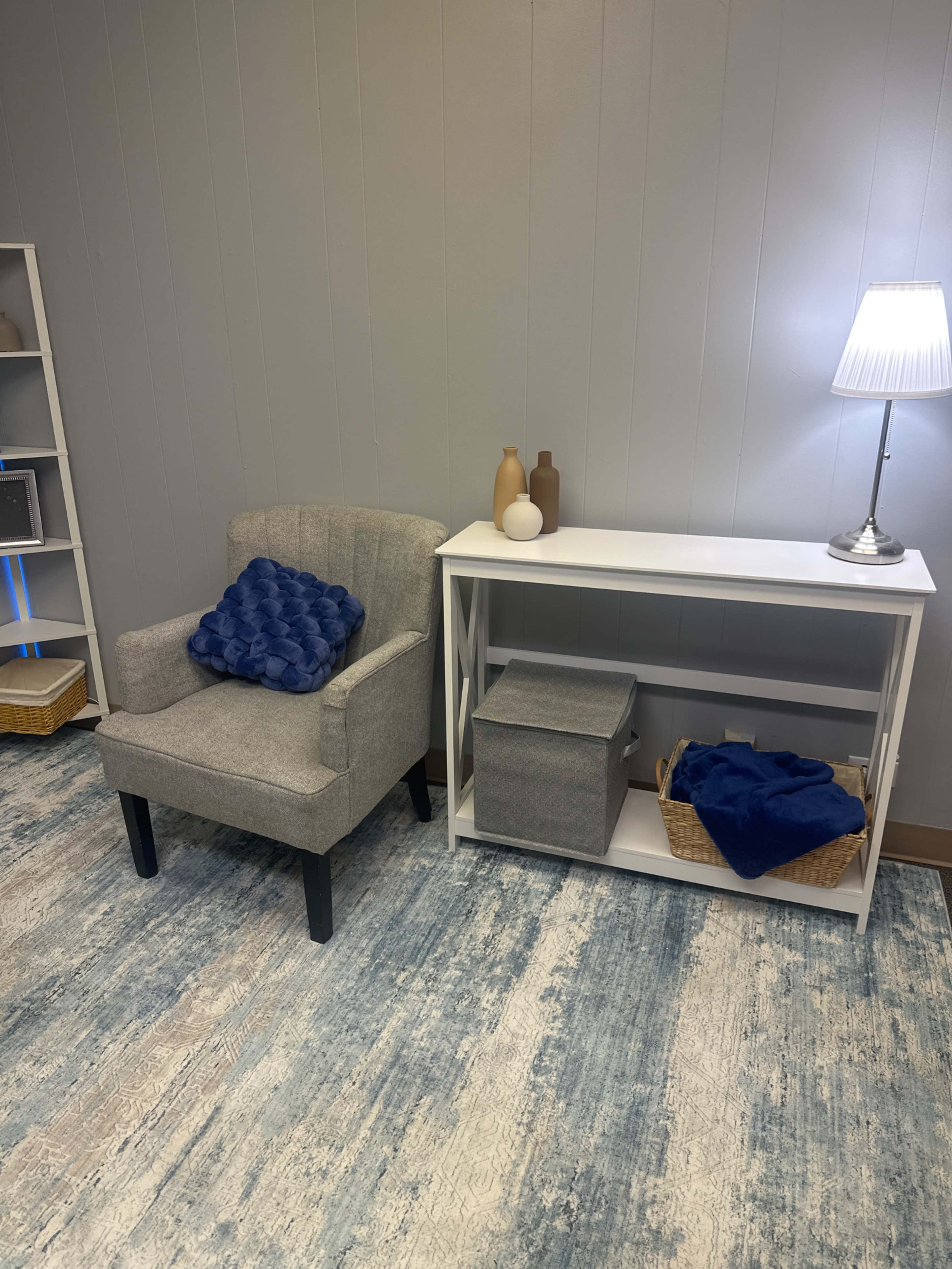 A gray armchair sits next to a white console table with decorative items, while a lamp provides light in a simple room.