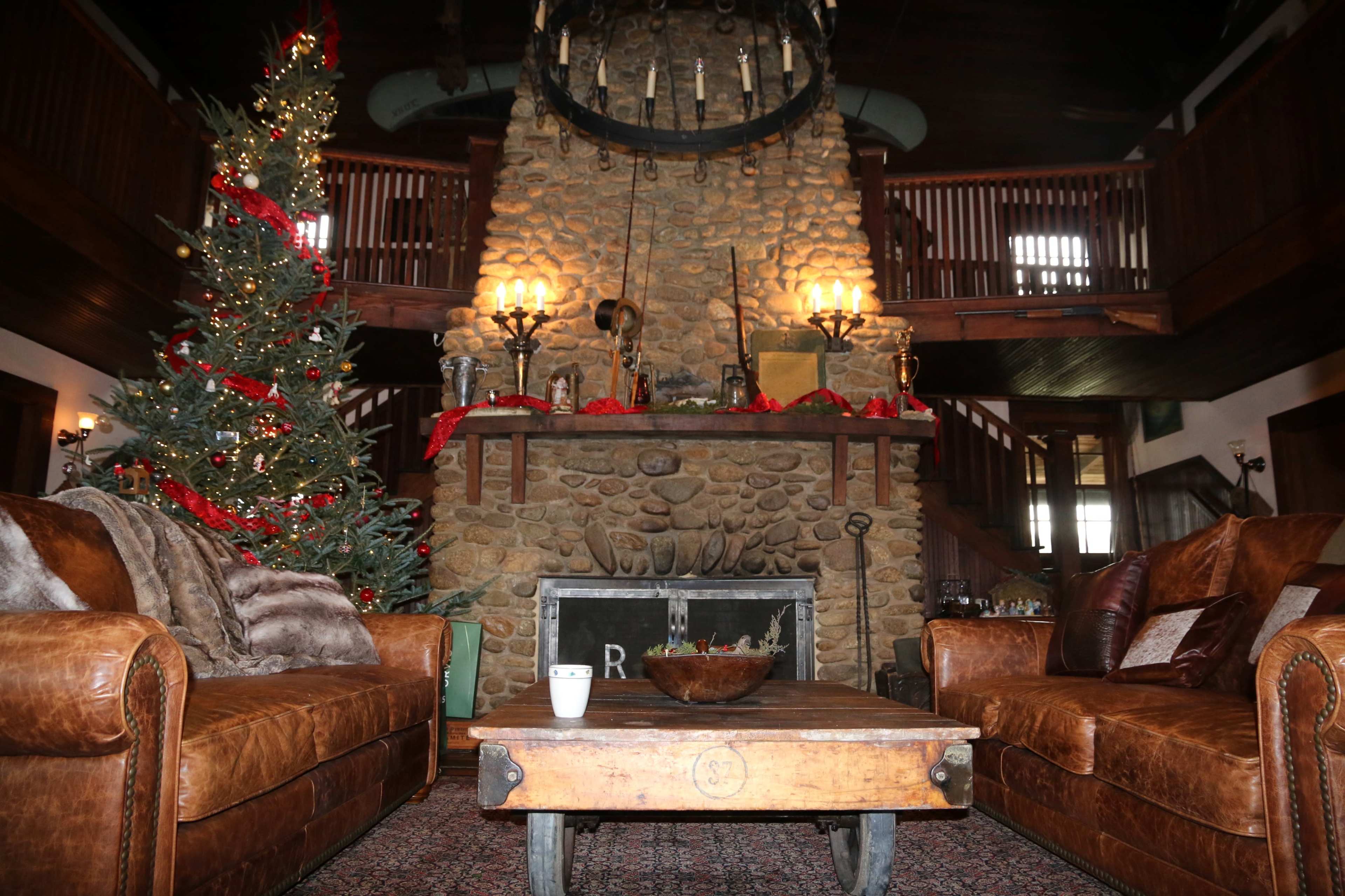 The image shows a cozy living room with a large stone fireplace, two brown leather sofas, a wooden coffee table, and a decorated Christmas tree.