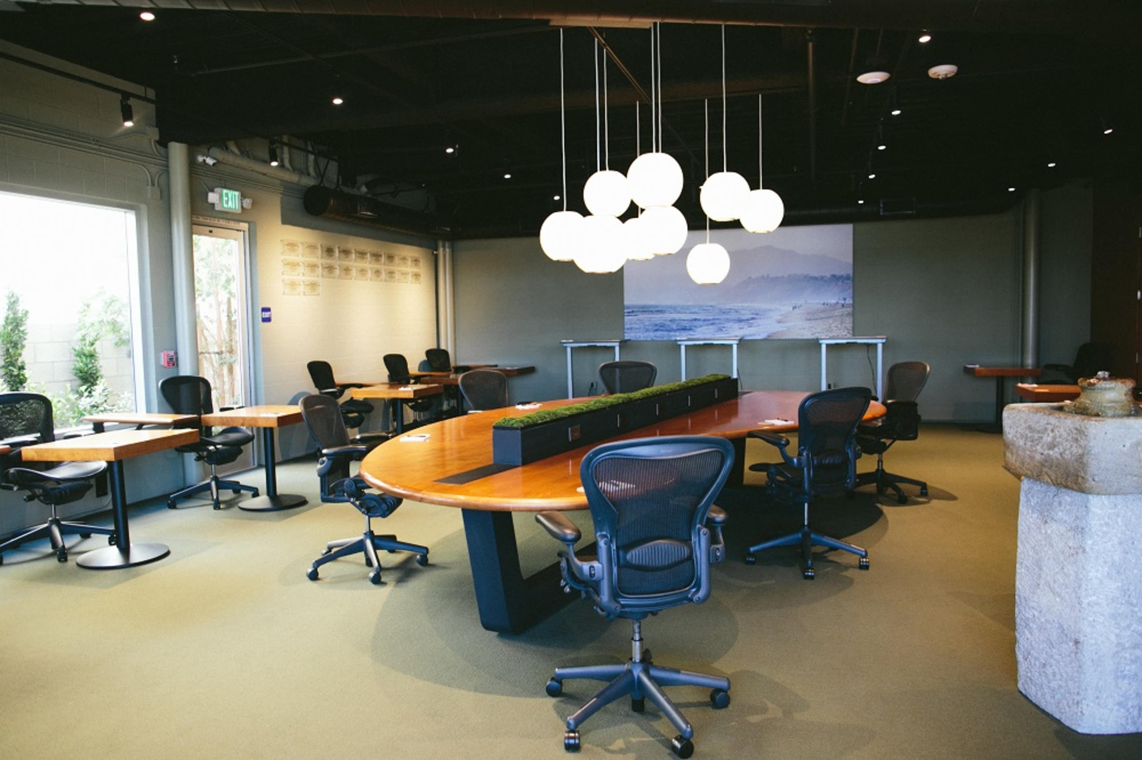 A modern meeting room features a large oval table surrounded by ergonomic chairs, with pendant lighting above and a scenic mural on one wall.