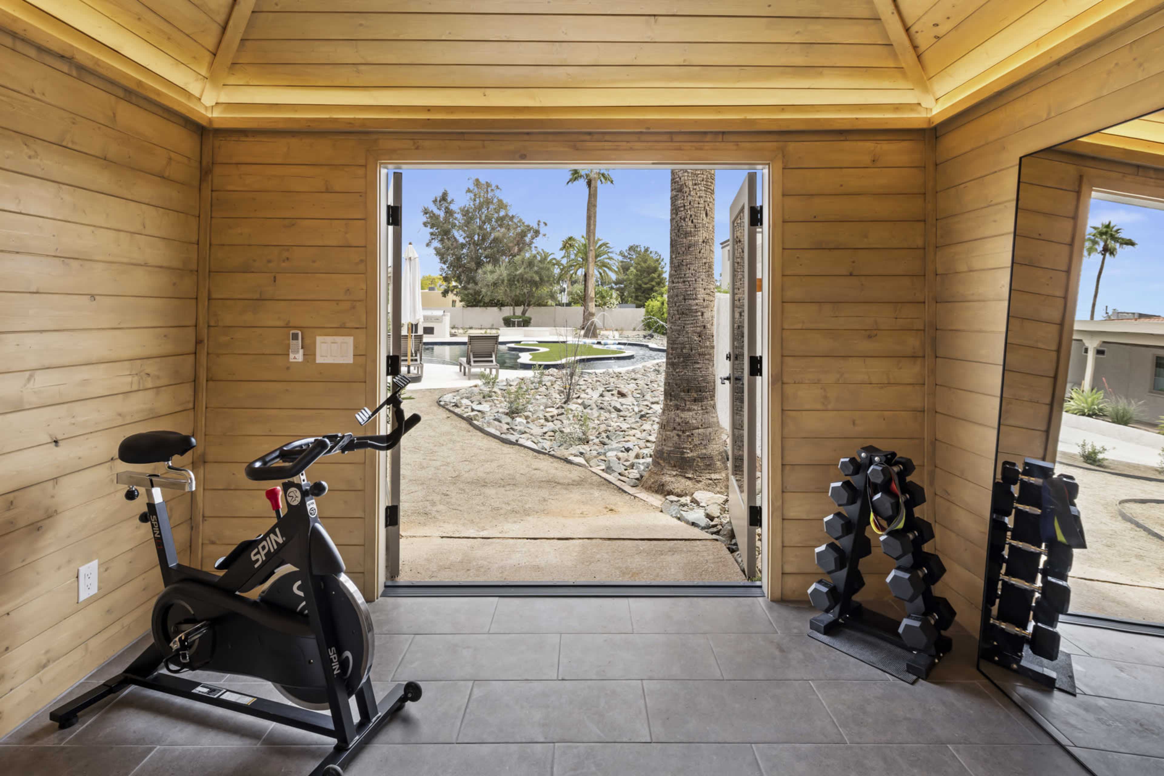 A well-lit indoor space features a stationary bike and weights, with a view of a landscaped outdoor area beyond a doorway.