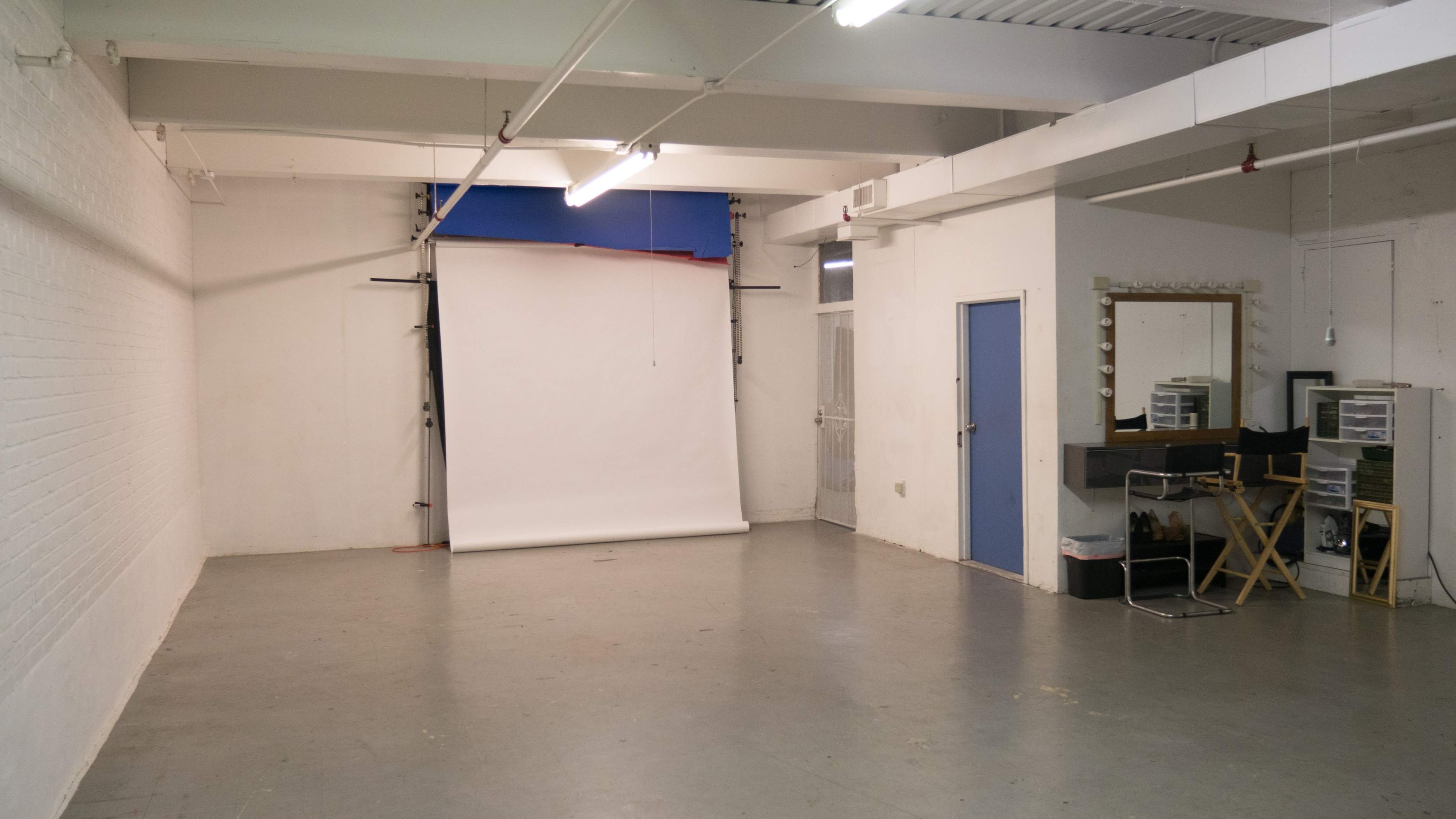 A sparsely furnished studio space with a white backdrop, a mirror, and a director's chair against a white brick wall.