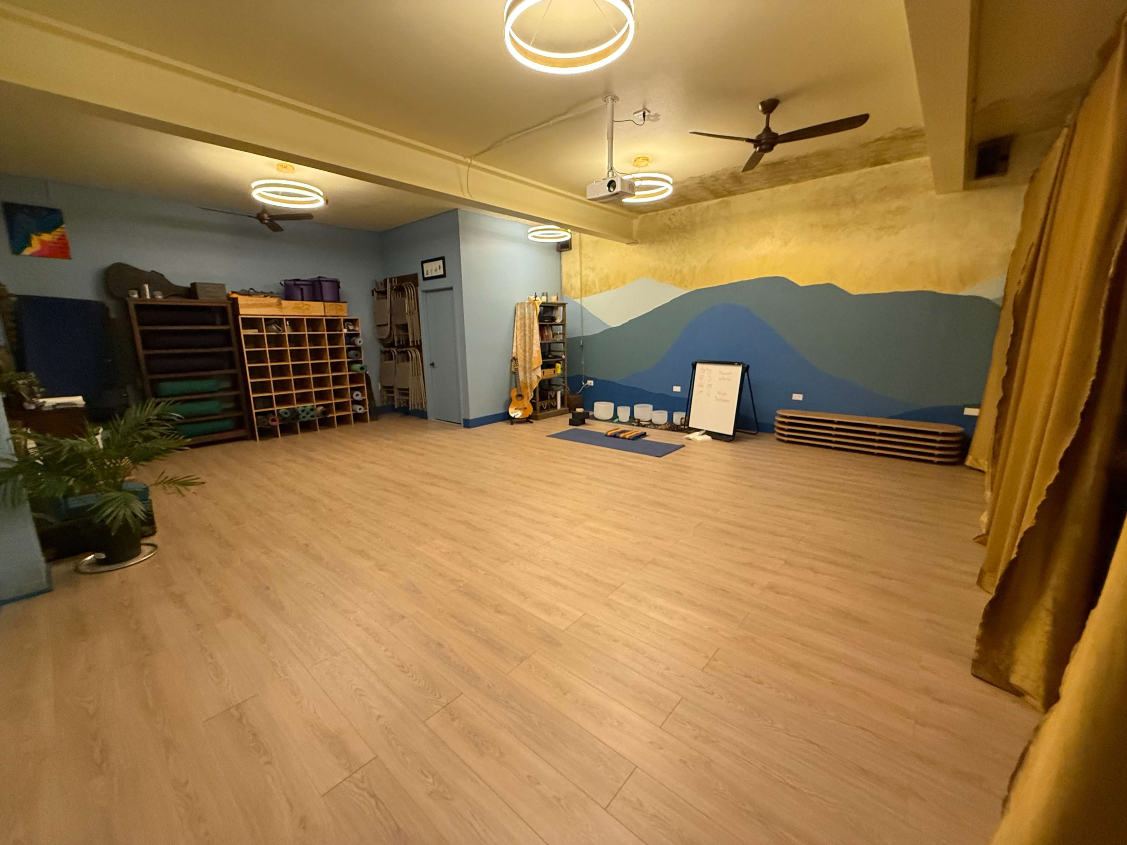The image shows a spacious yoga studio with wooden flooring, light blue walls featuring a mountain mural, and circular light fixtures hanging from the ceiling.