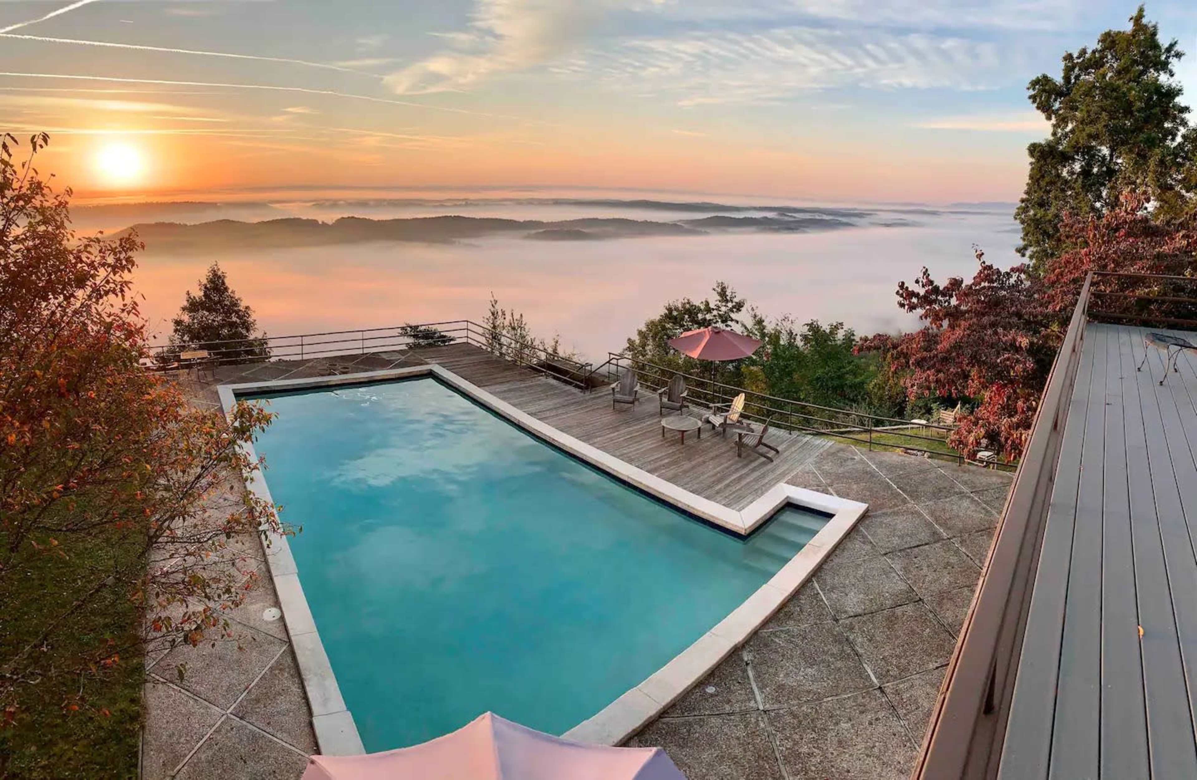 A rectangular swimming pool is surrounded by a wooden deck, overlooking a landscape of misty hills at sunrise.