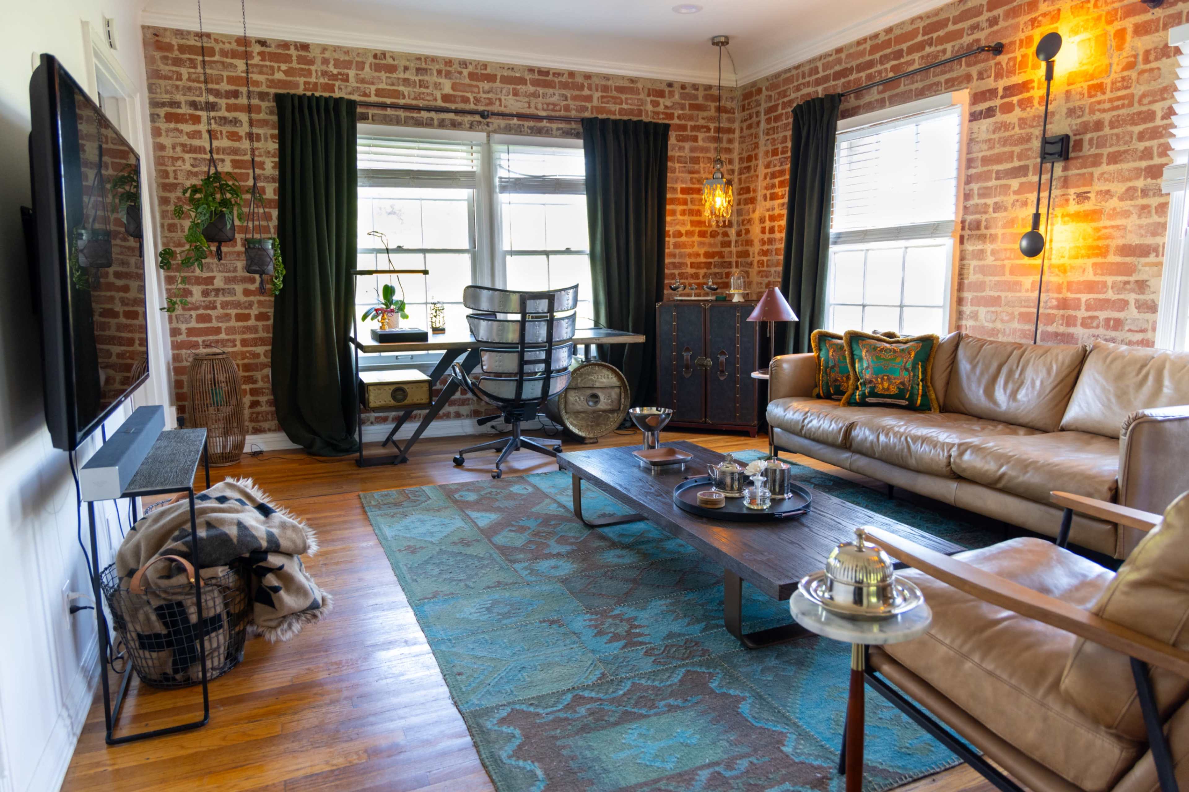 The image shows a cozy living room with brick walls, hardwood flooring, a leather sofa, a coffee table, and a workspace with a chair and desk by a window.