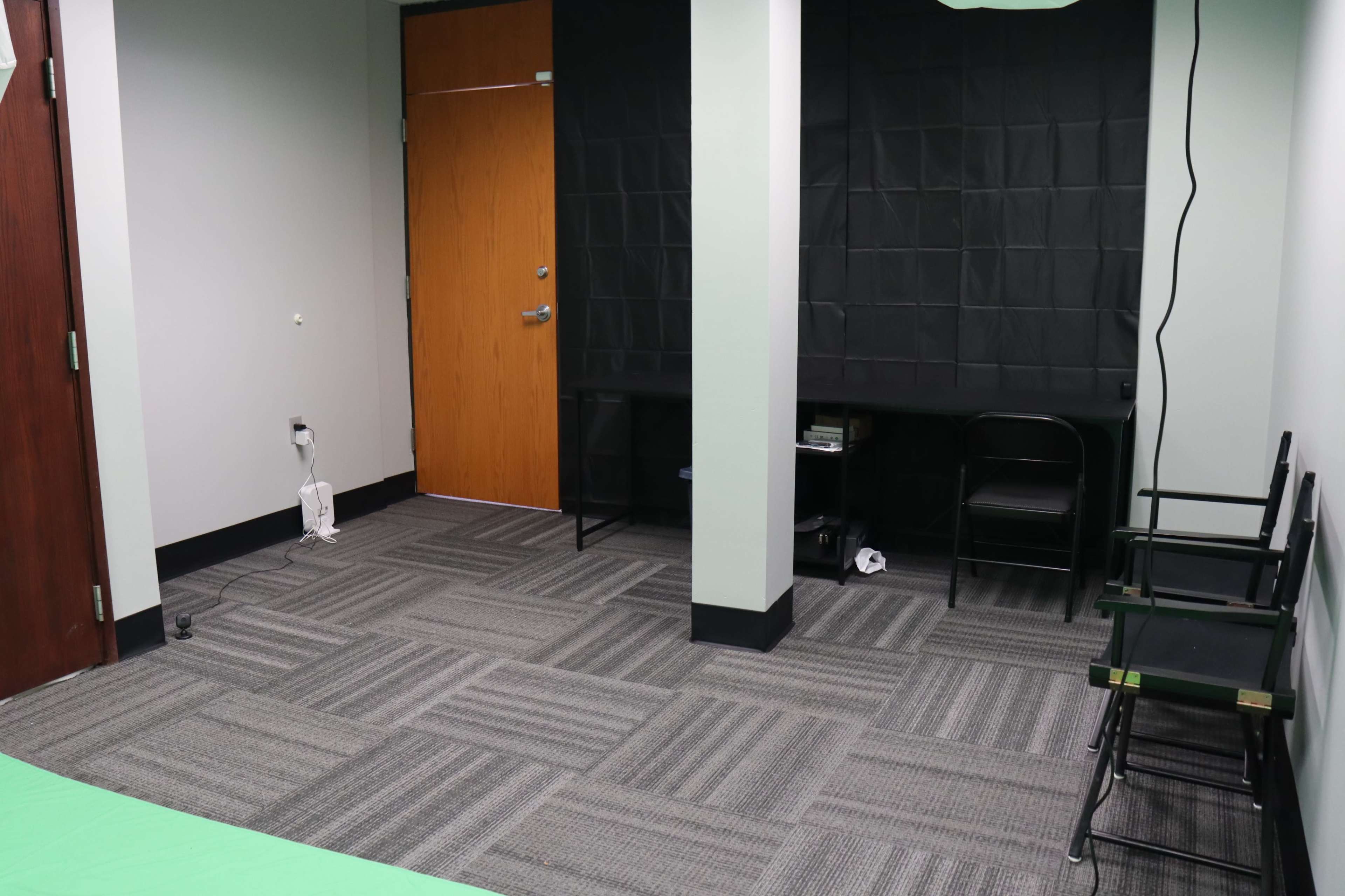 The image shows a minimalistic room with gray carpeting, light green walls, and two black chairs placed next to a dark backdrop.