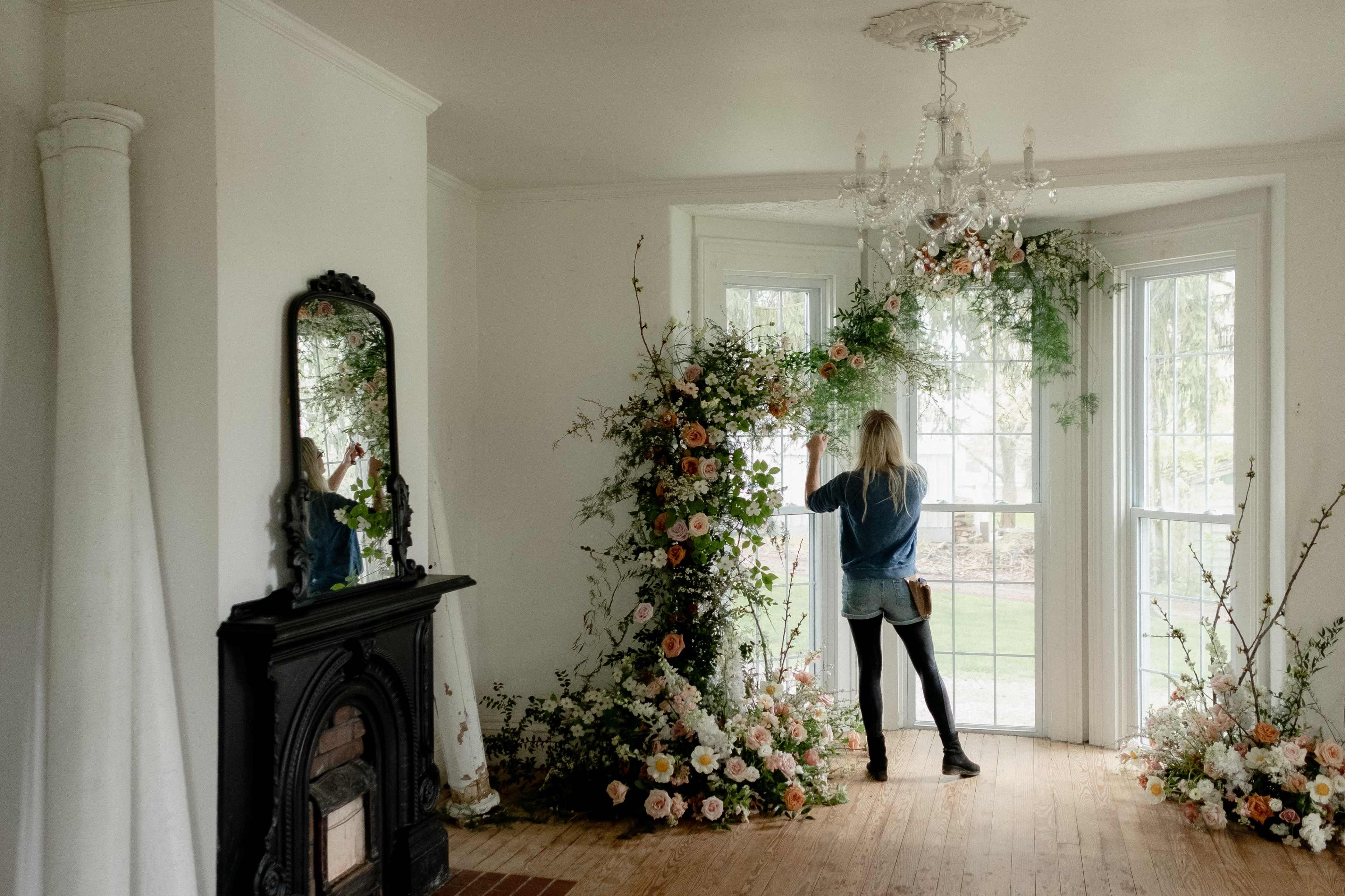 A woman stands by a large window adorned with an array of flowers, while a mirror reflects part of the room and a chandelier hangs overhead.