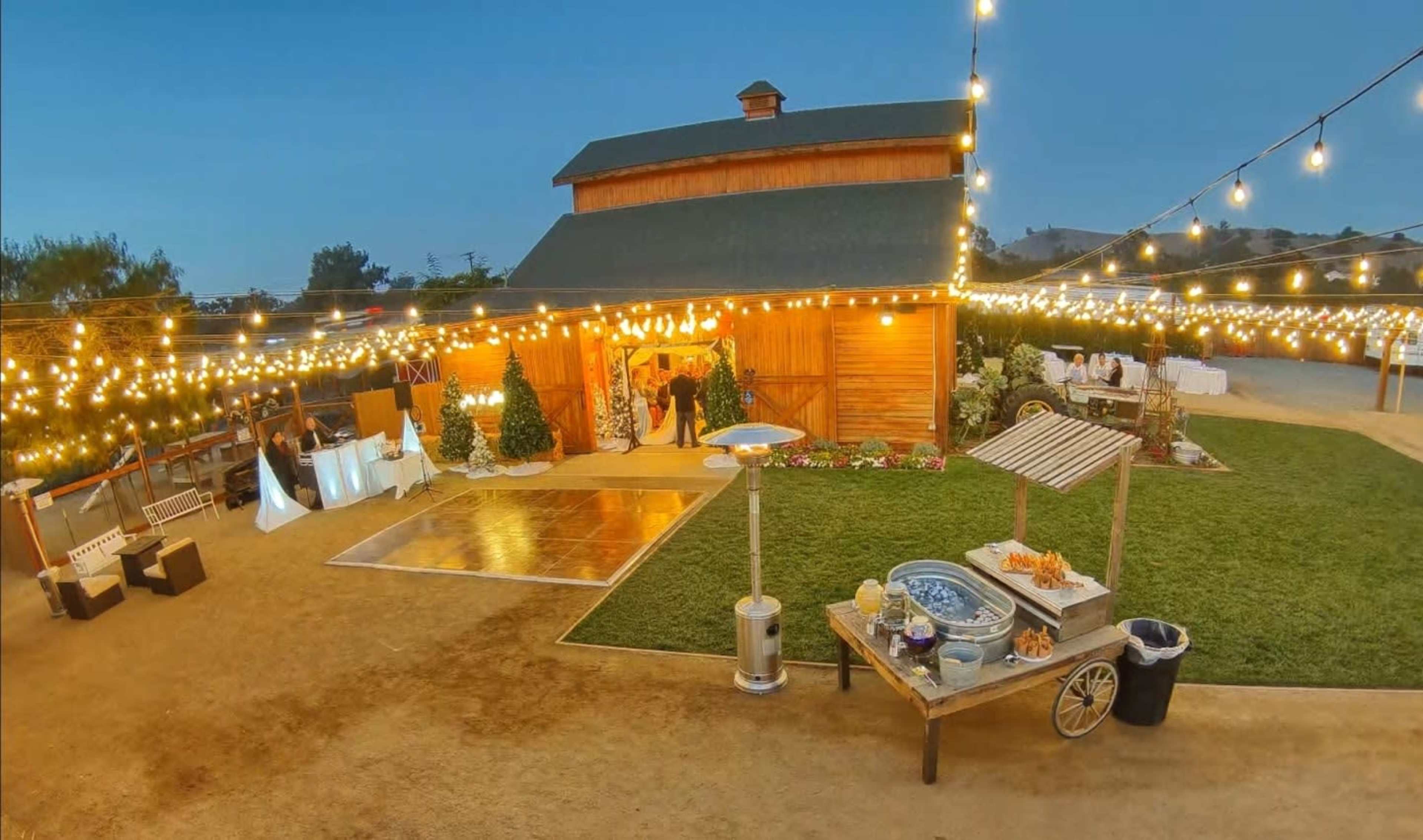 The image shows a barn-style venue illuminated by string lights, with outdoor seating, a dance floor, and a refreshment cart in a landscaped area.