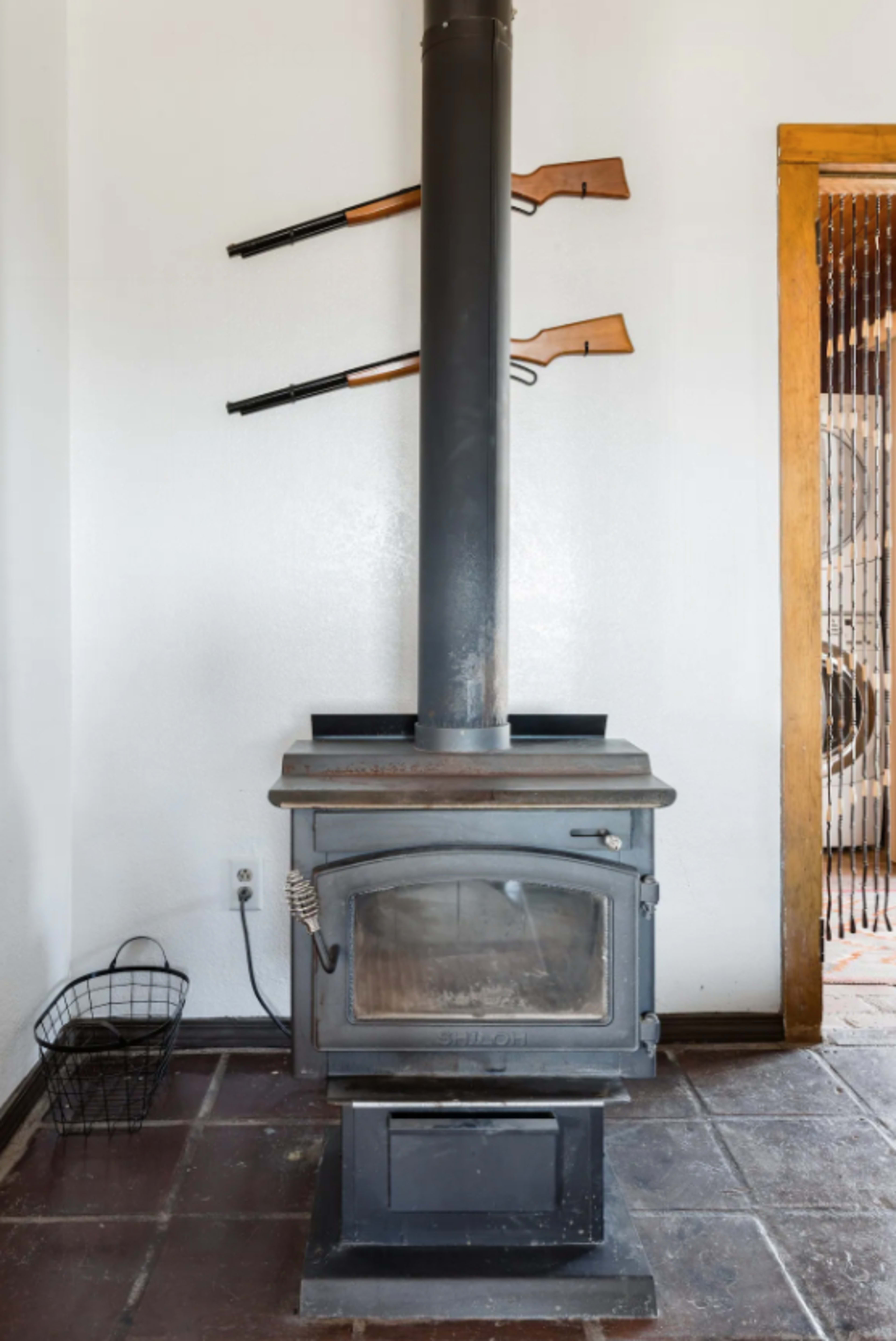 A metal wood-burning stove stands against a white wall, with two rifles mounted above it and a wire basket nearby.