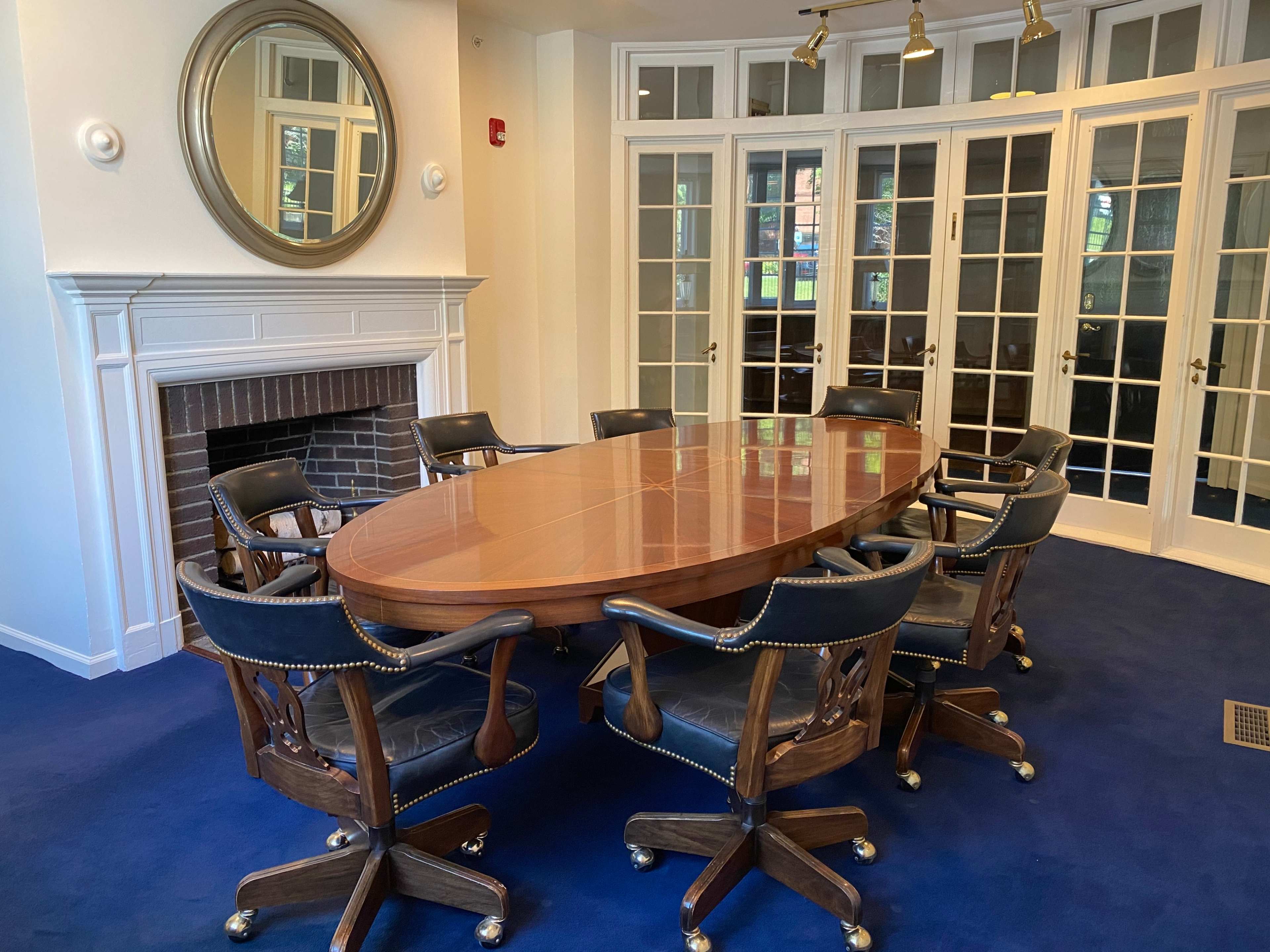 A conference room features an oval wooden table surrounded by eight black leather rolling chairs, with a large mirror above a fireplace and glass doors leading to an outside view.