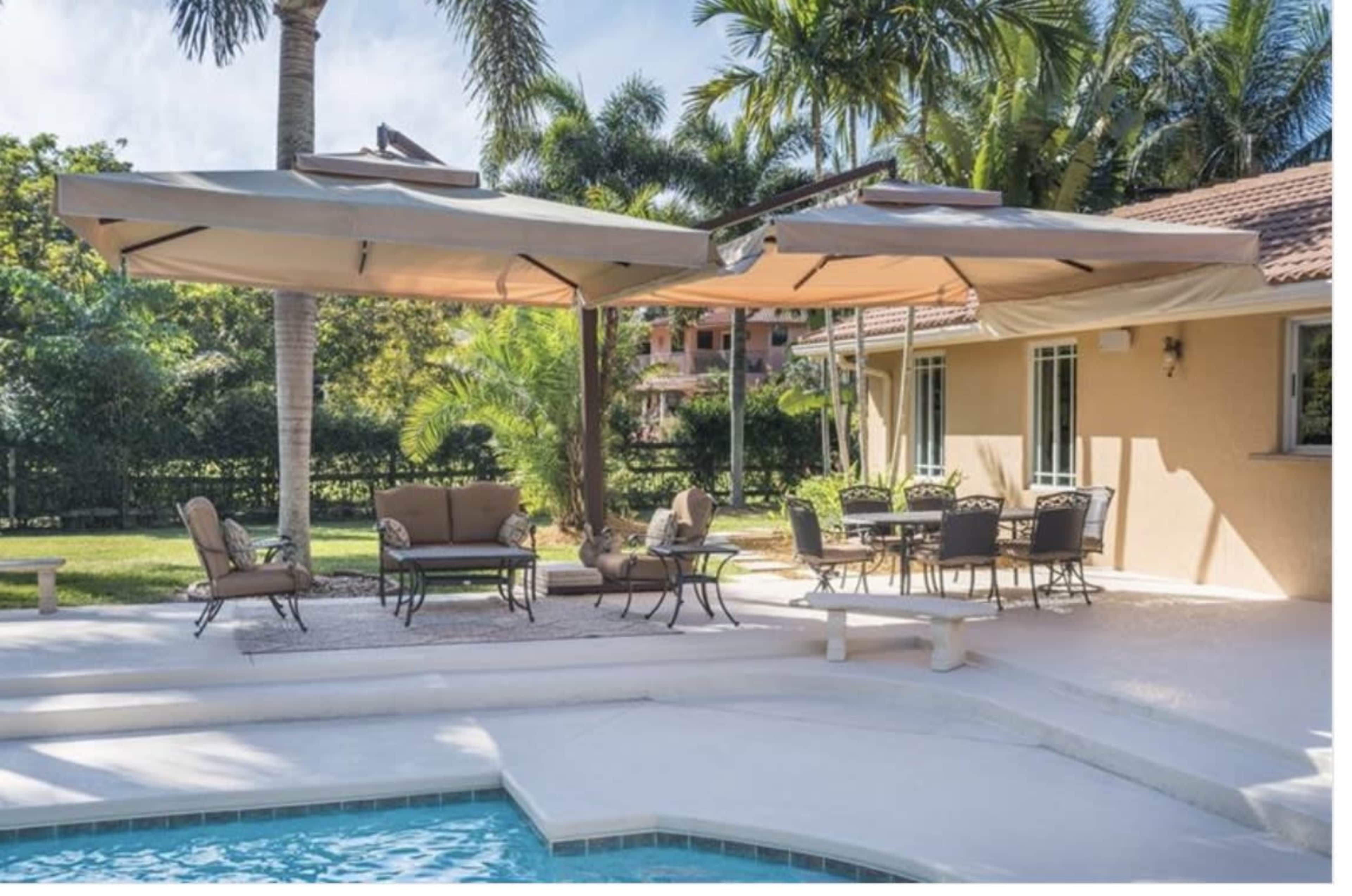 A patio area with a pool features a seating arrangement under large umbrellas and a dining table surrounded by palm trees.