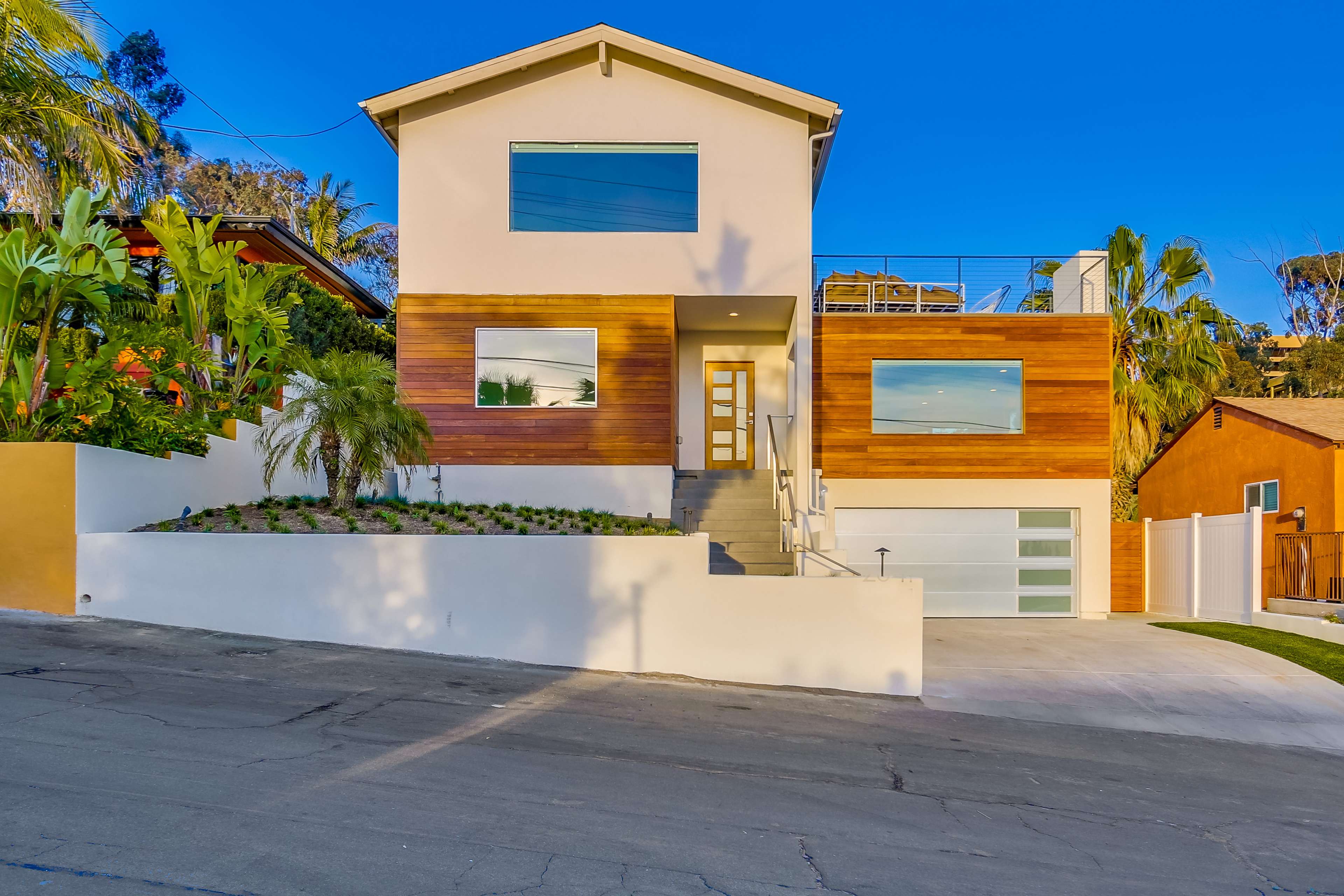 The image shows a modern two-story house with a mix of white and wooden exterior, large windows, and a landscaped front yard on a sloped street.