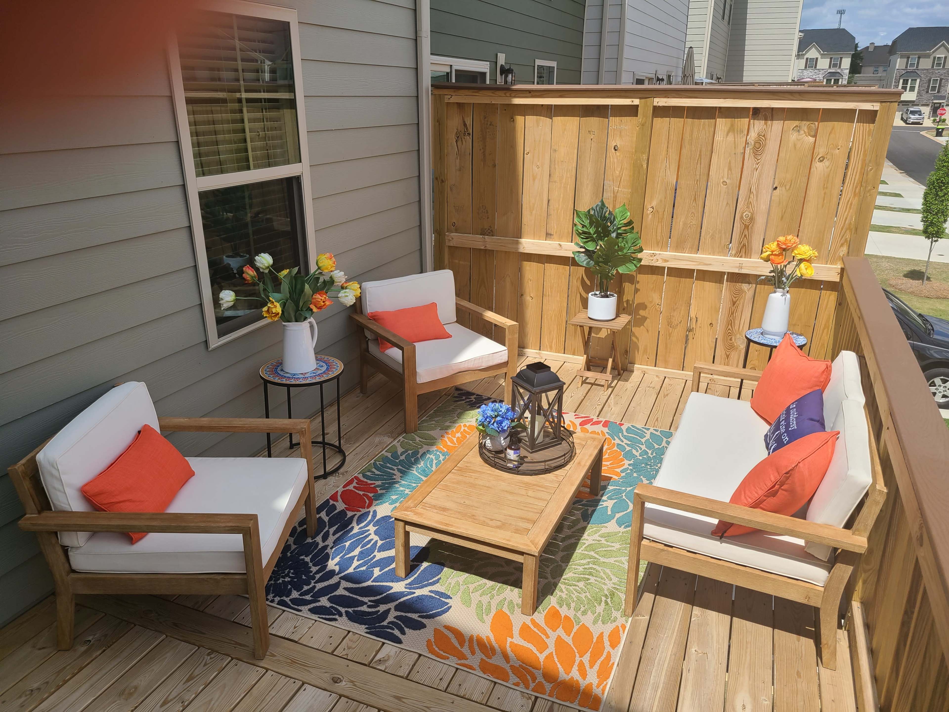 A small outdoor balcony with two armchairs, a coffee table, a colorful rug, and decorative plants surrounded by wooden fencing.