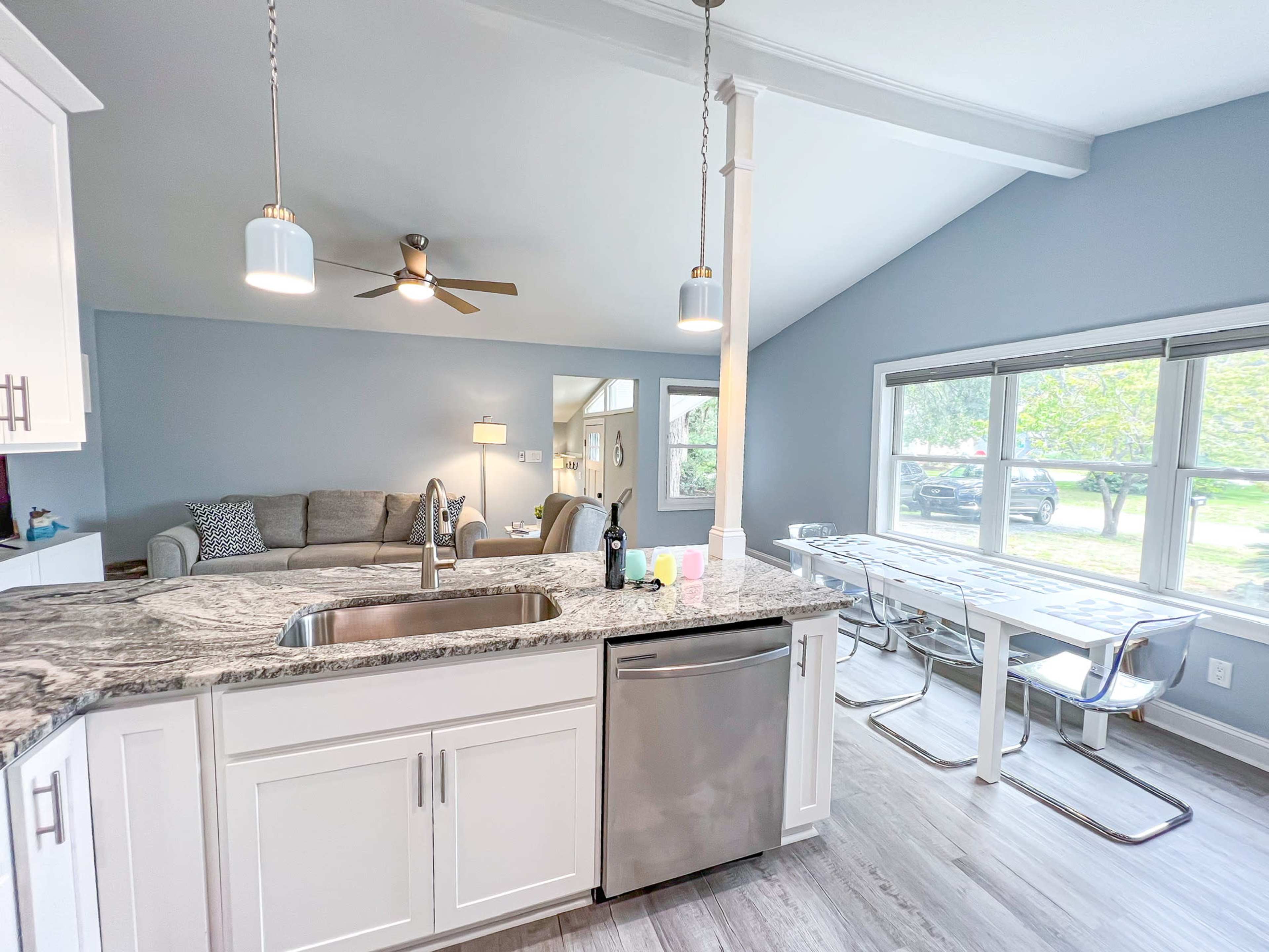 A modern kitchen with white cabinets, a marble countertop, a stainless steel sink, and a living room in the background featuring a gray sofa and large windows.