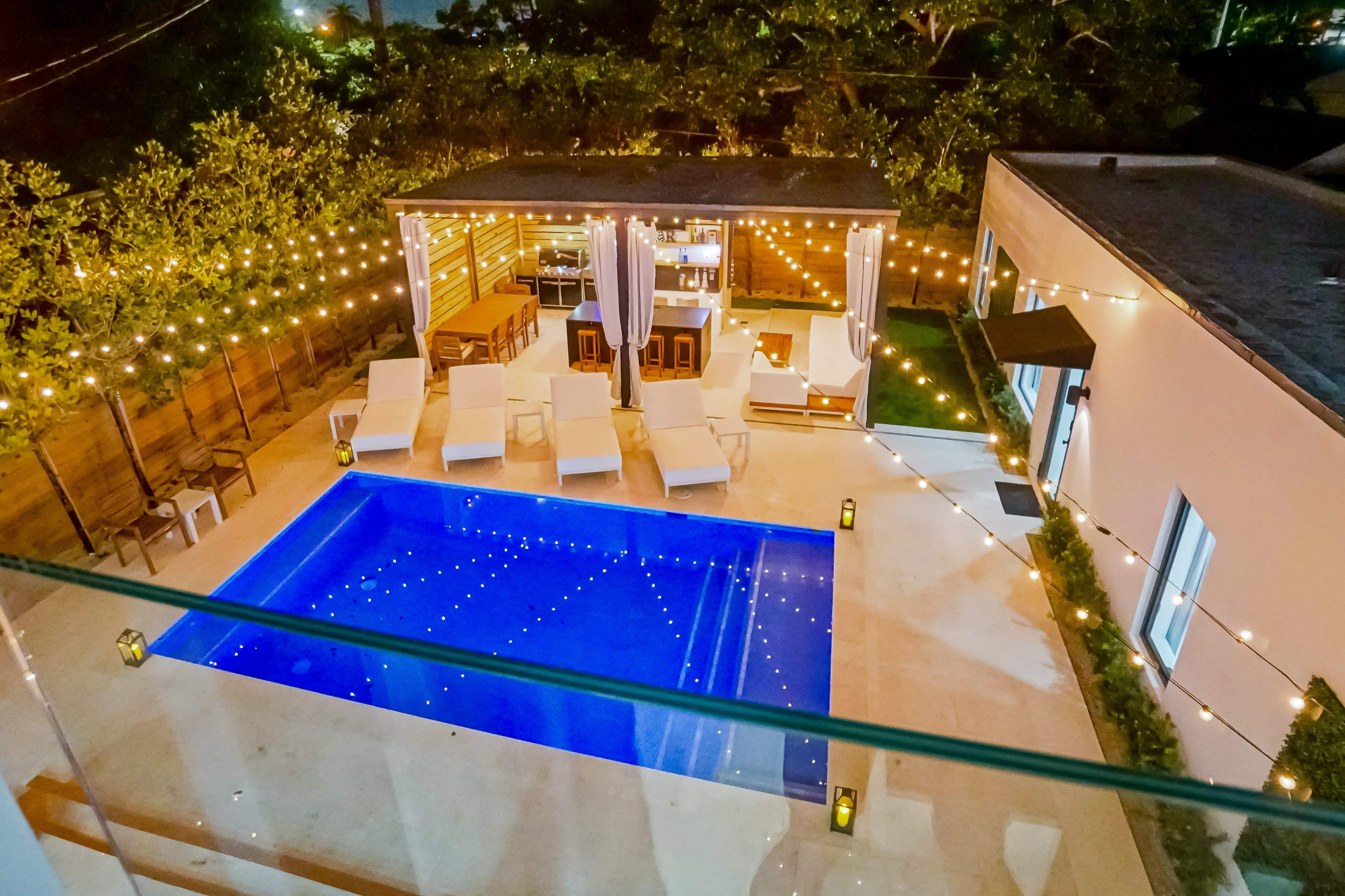 The image shows an outdoor area featuring a clear swimming pool surrounded by lounge chairs, with string lights illuminating the space and a covered seating area off to the side.