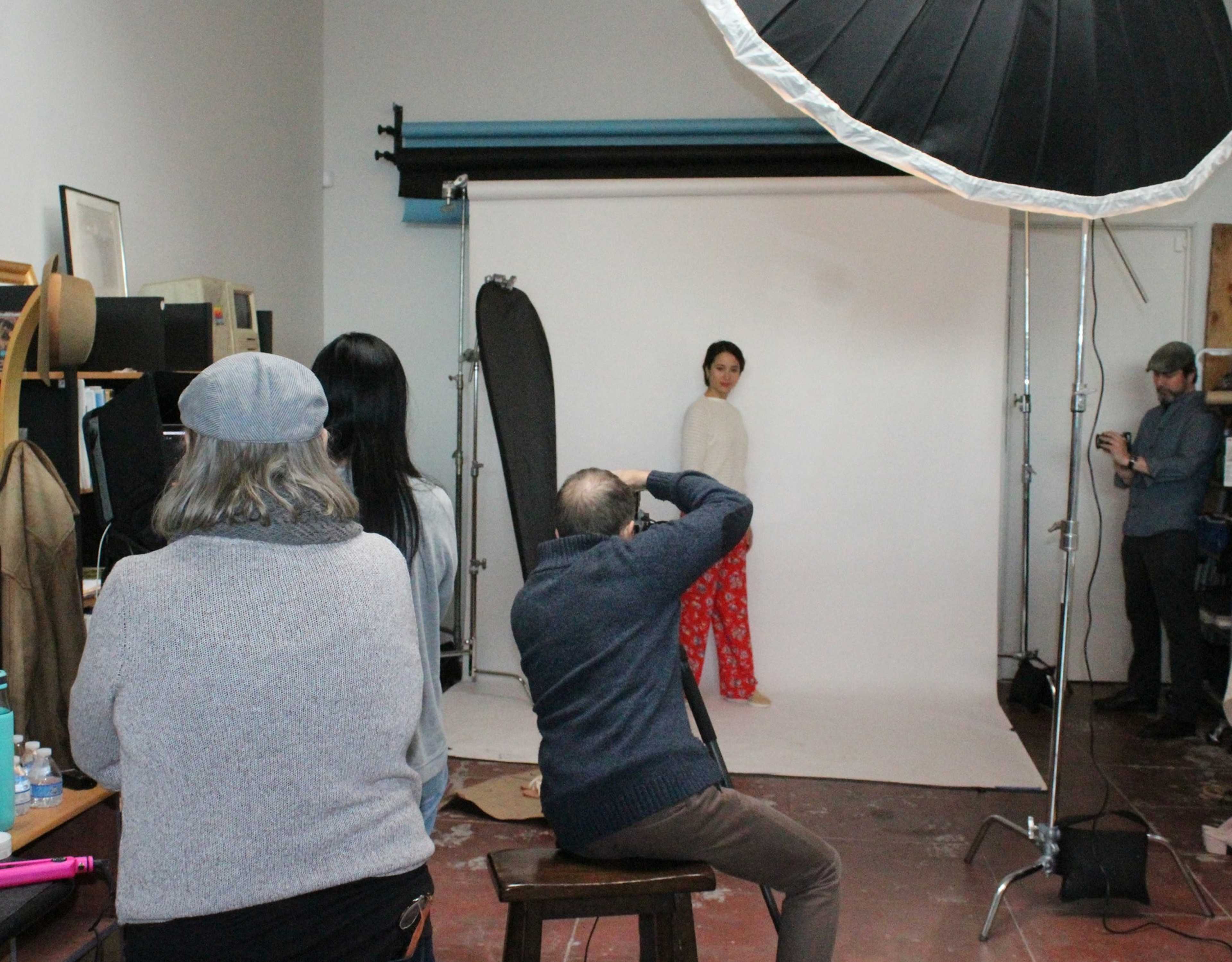 A model poses in front of a professional backdrop while a photographer and a small group of onlookers observe in a studio setting.