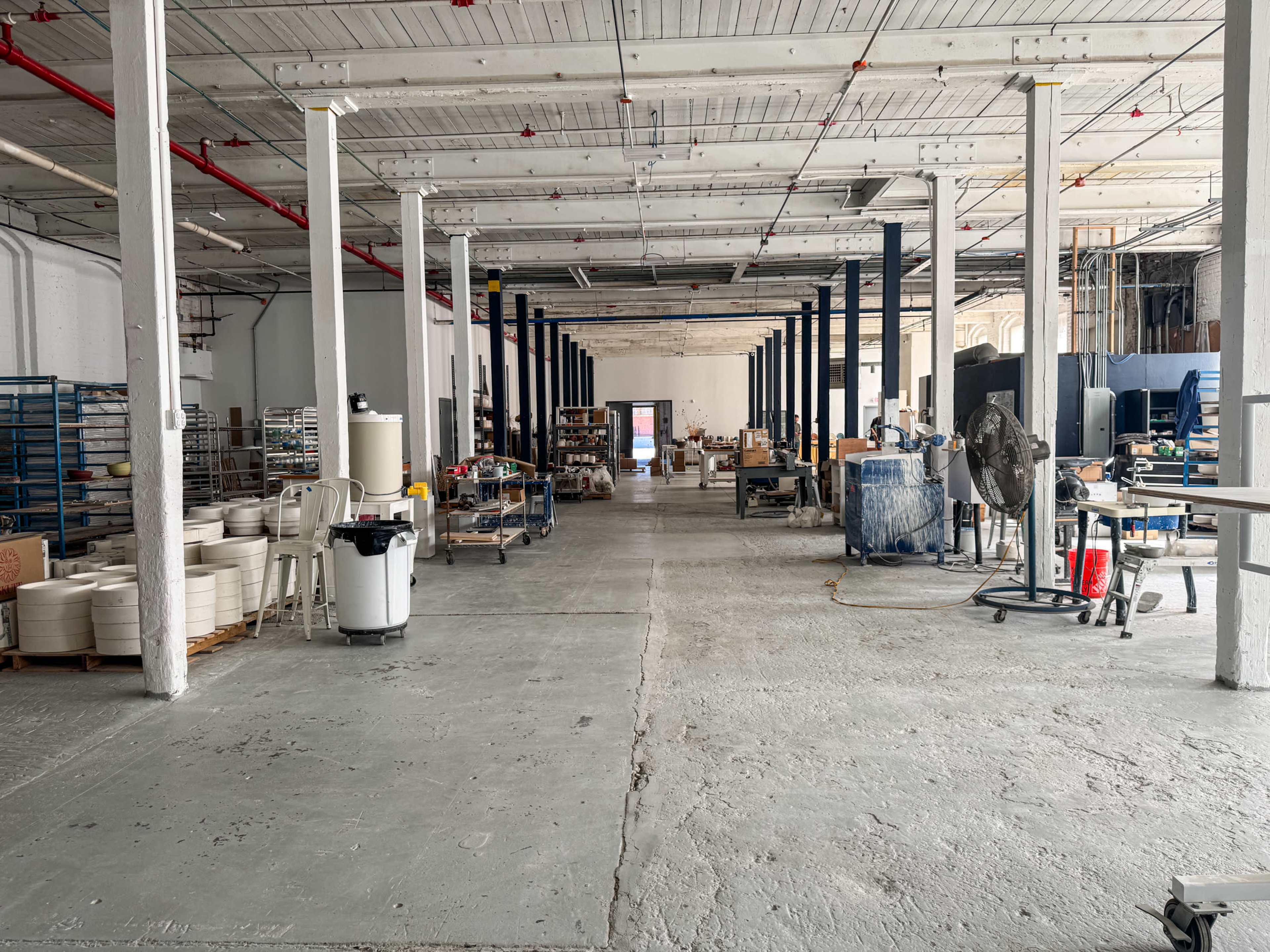 The image shows a spacious, empty industrial warehouse with rows of tables and equipment along the sides, and a long aisle leading to an open doorway at the far end.