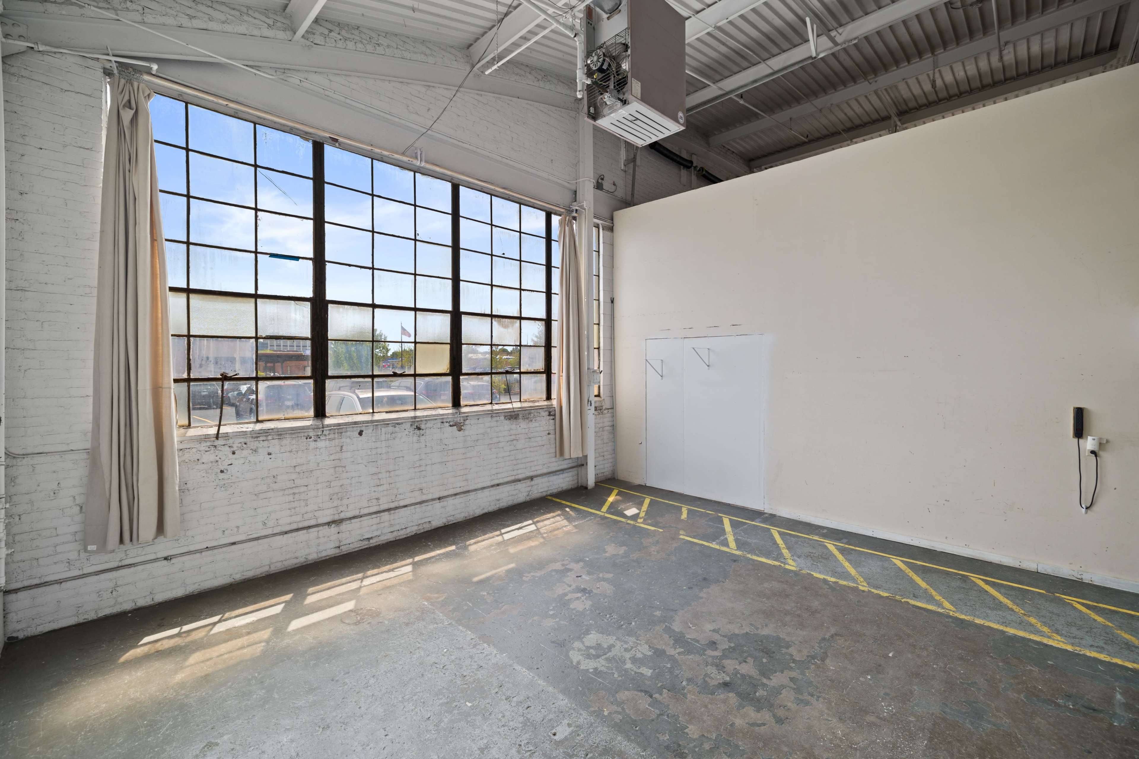 A spacious, light-filled room featuring large industrial windows, bare white walls, and a concrete floor marked with yellow lines.