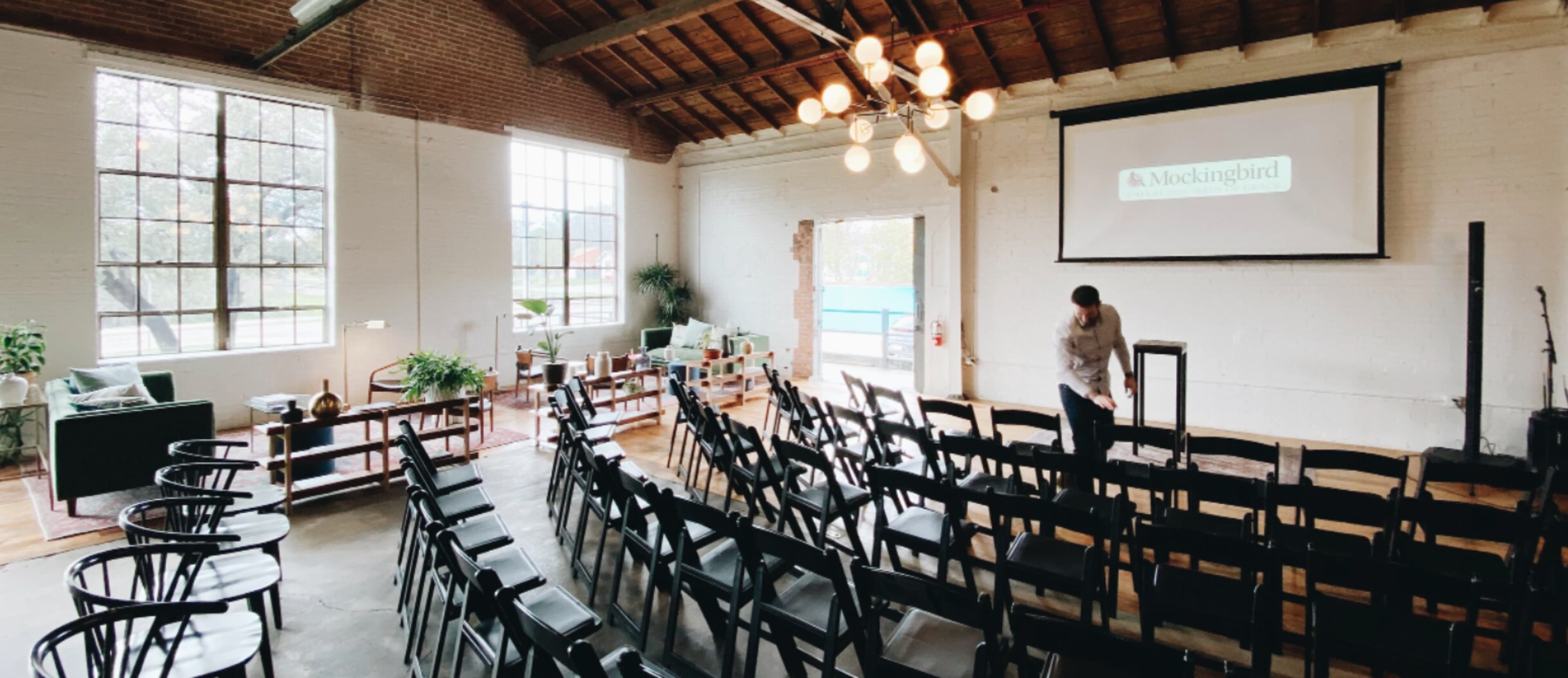 A spacious event room with rows of black chairs facing a presentation screen, surrounded by large windows and modern furniture.