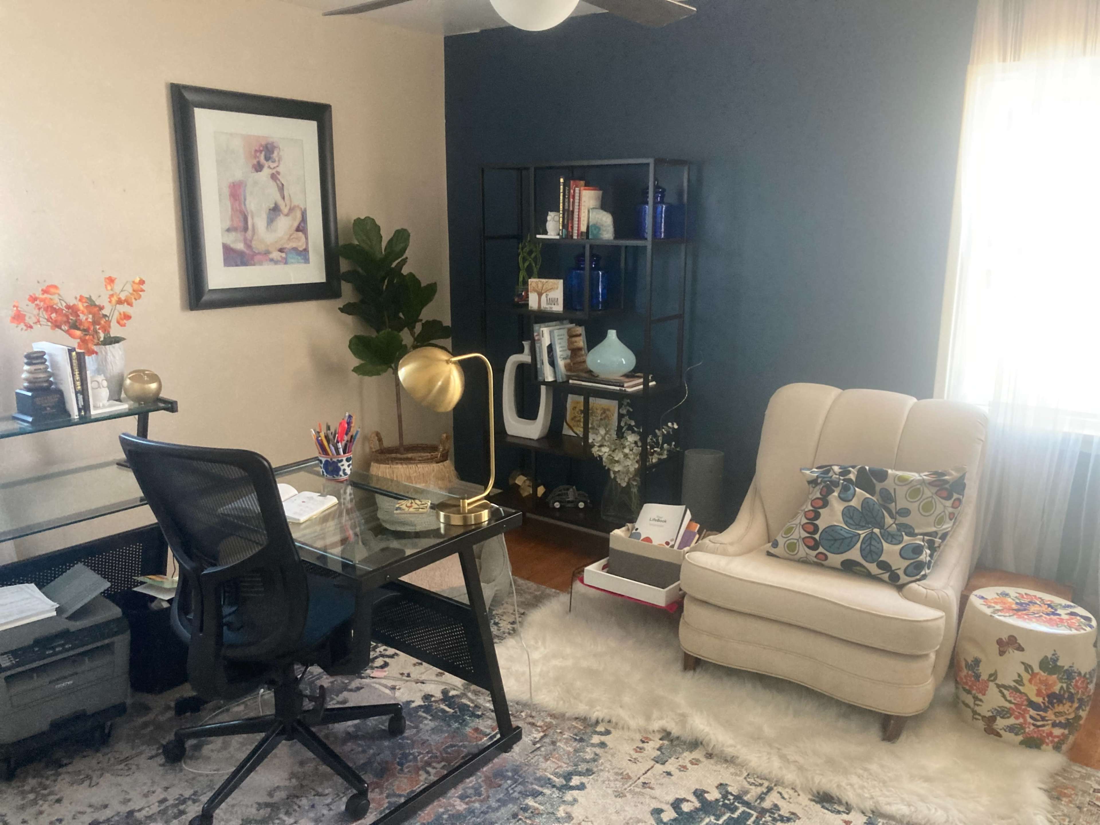 A home office space with a glass desk, a stylish armchair, a shelving unit, a plant, and decorative items on a patterned rug.