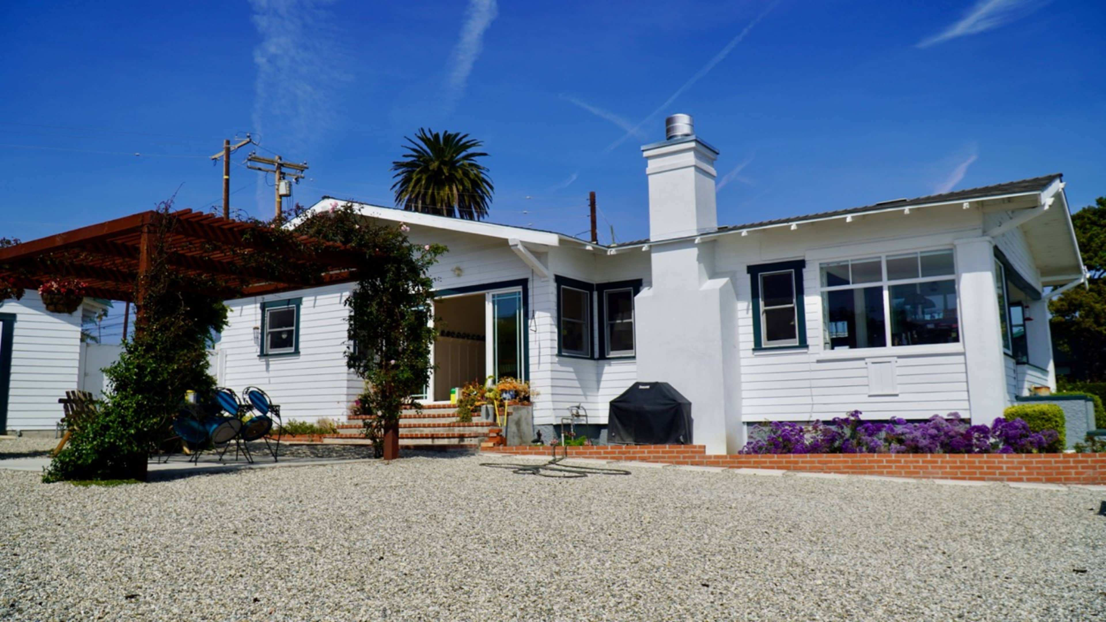 A white wooden house with a front porch, surrounded by gravel and featuring a pergola with seating and colorful flowers.