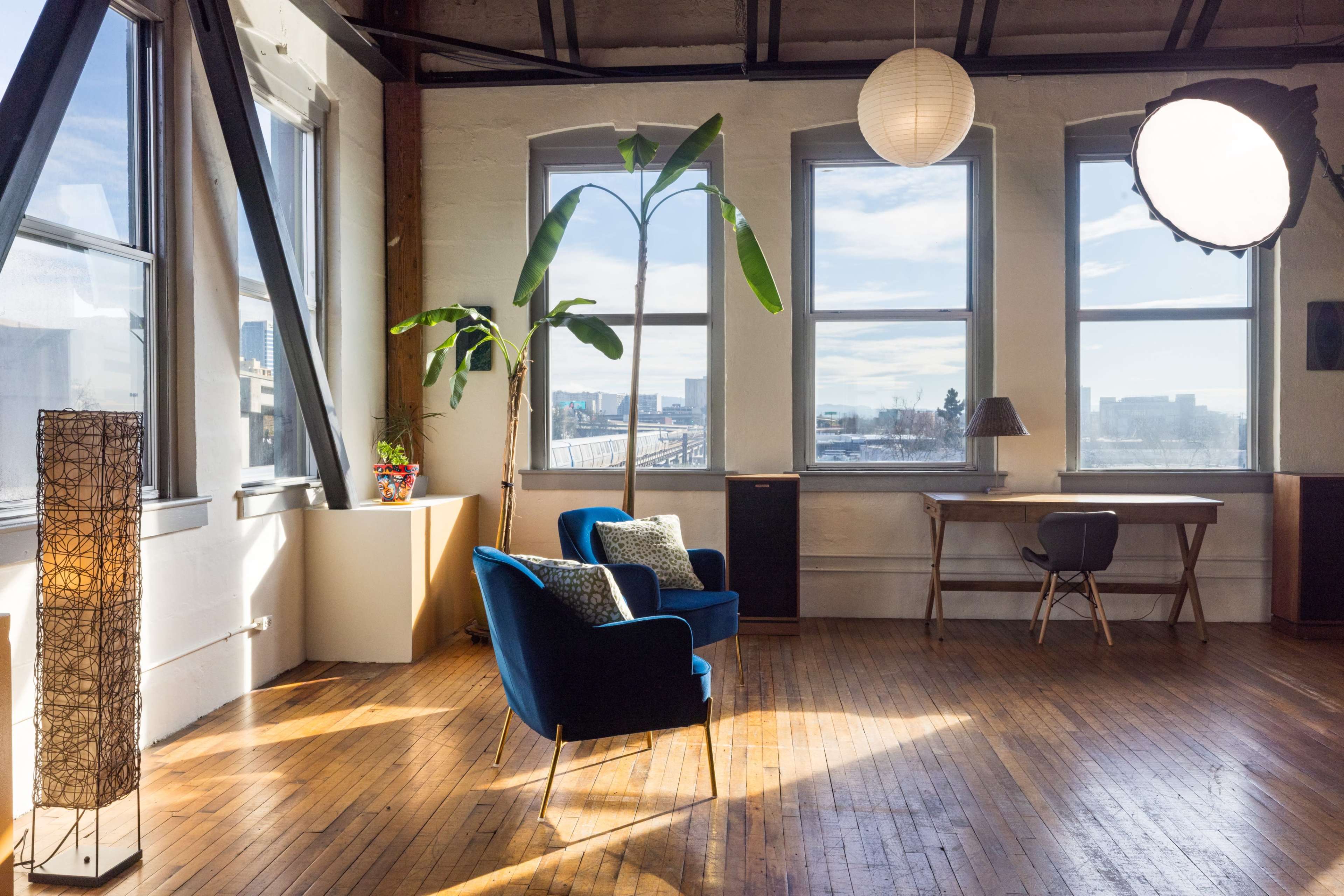 The image shows a bright, spacious room with large windows, featuring two blue chairs, a desk, a plant, and a unique light fixture.
