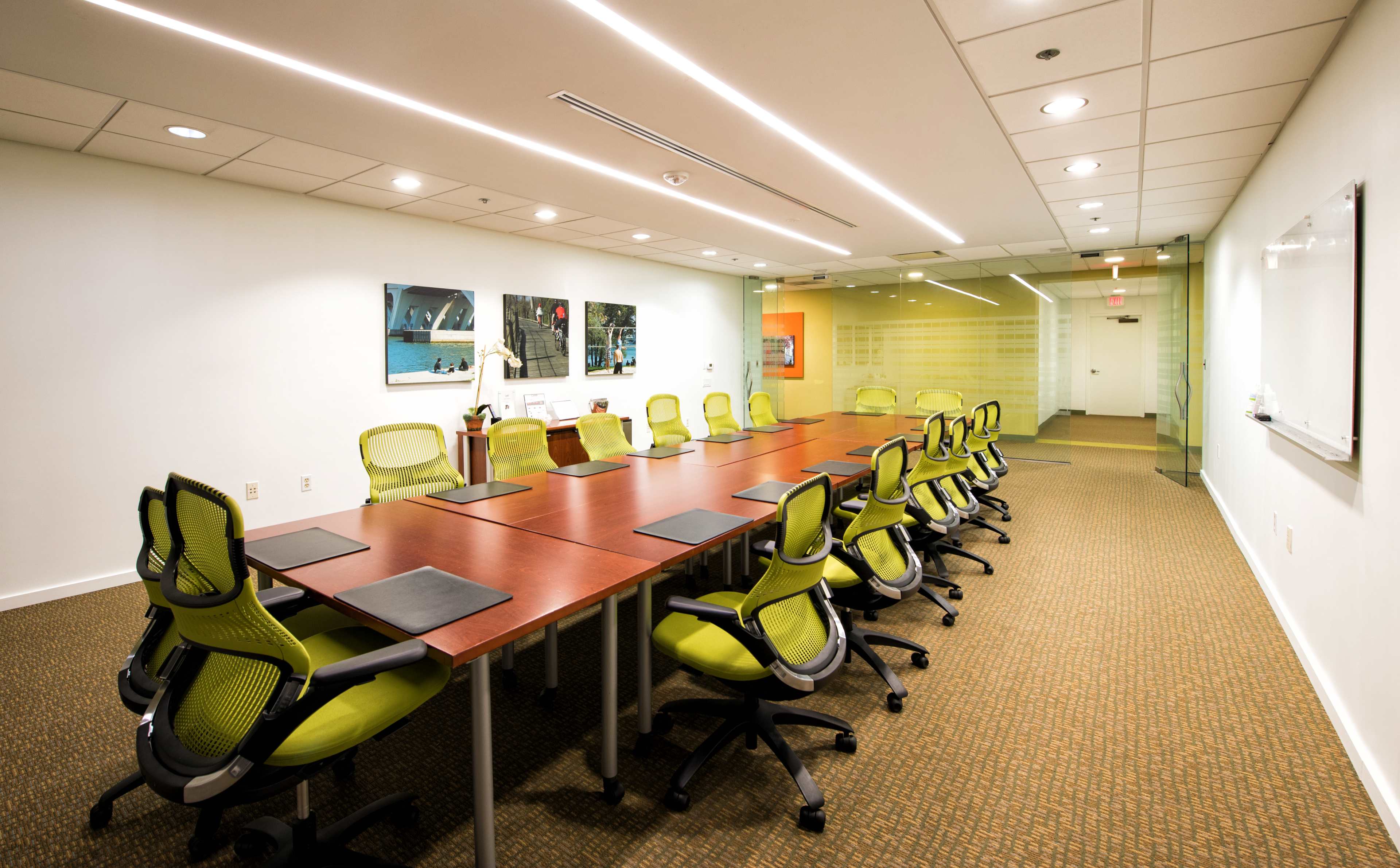 A modern conference room features a long wooden table surrounded by twelve green chairs, with a whiteboard and glass doors visible in the background.