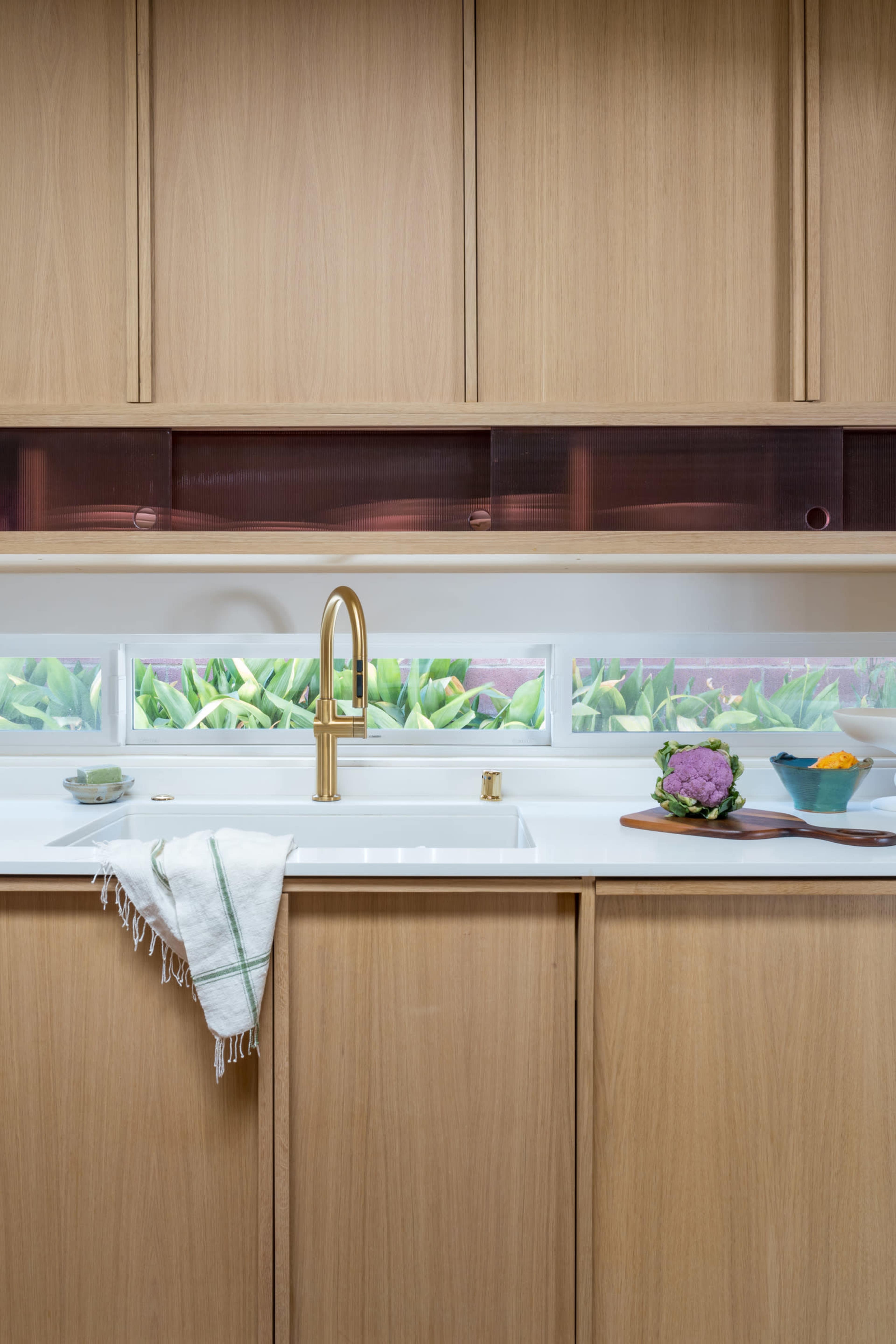 The image shows a modern kitchen with light wood cabinetry, a white sink, and a brass faucet, featuring a window that displays greenery outside.