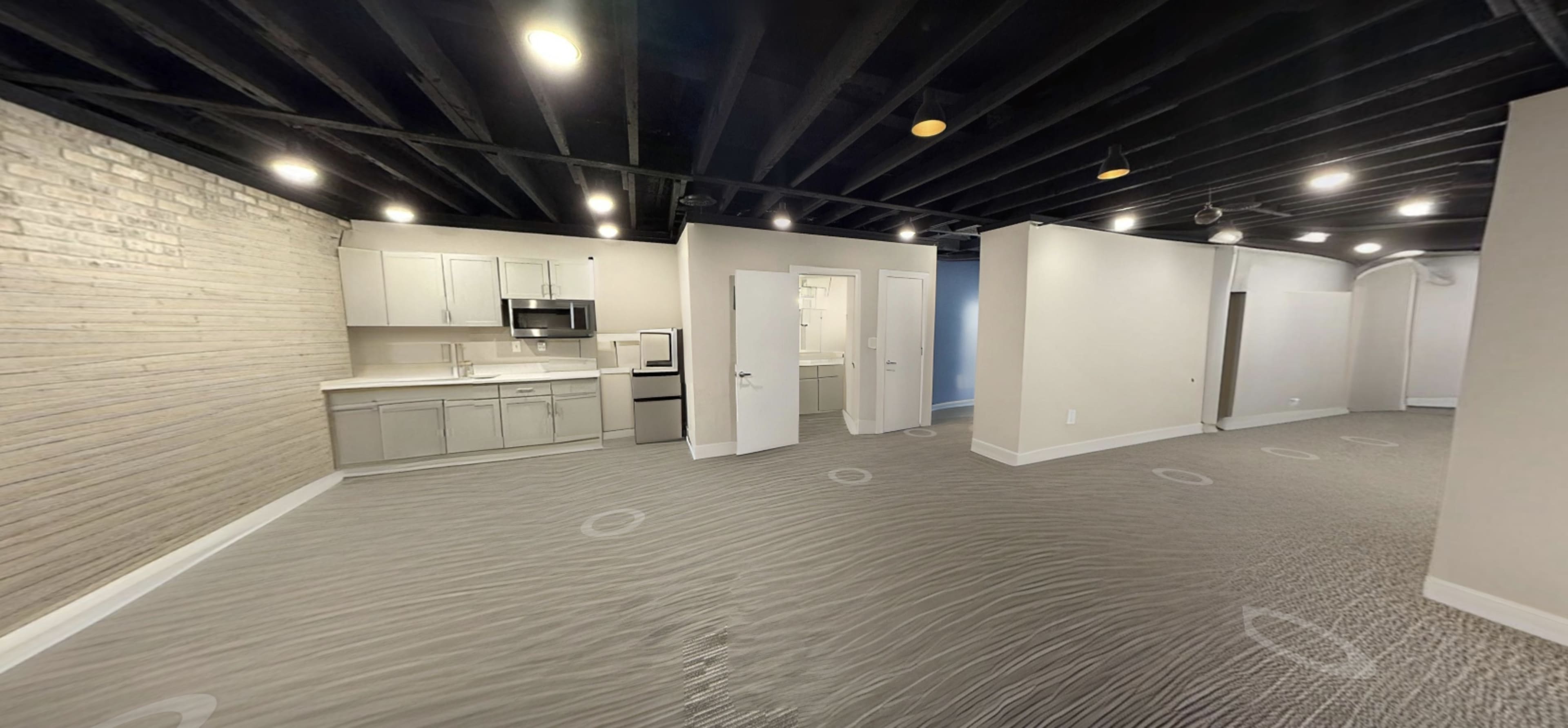 The image shows an open-concept living space featuring a kitchen area with modern appliances, light-colored cabinetry, and a large carpeted area.