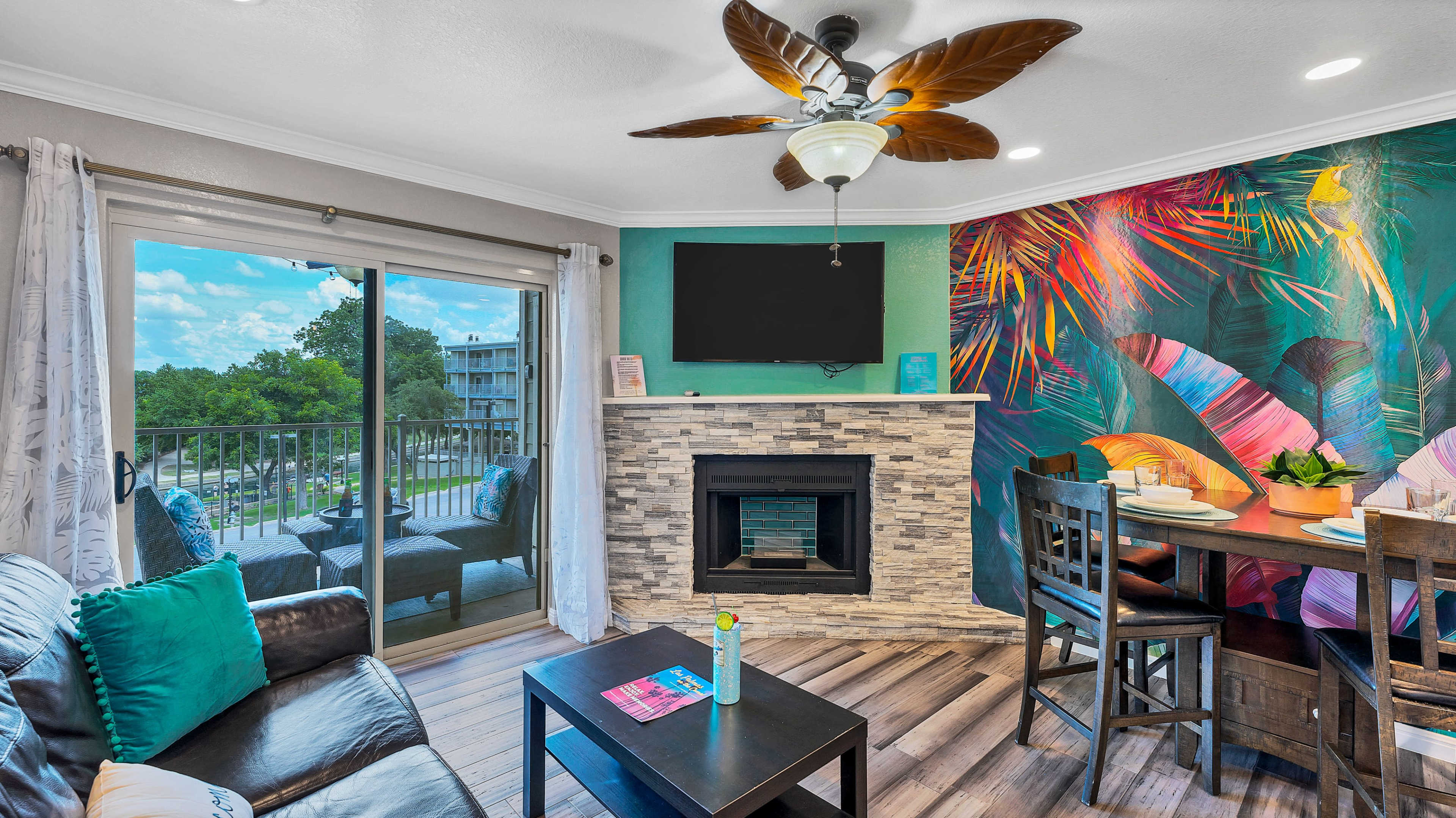 A living area featuring a dark leather sofa, a stone fireplace, a dining table, and a vibrant mural with tropical foliage, alongside a balcony door leading to an outdoor view.