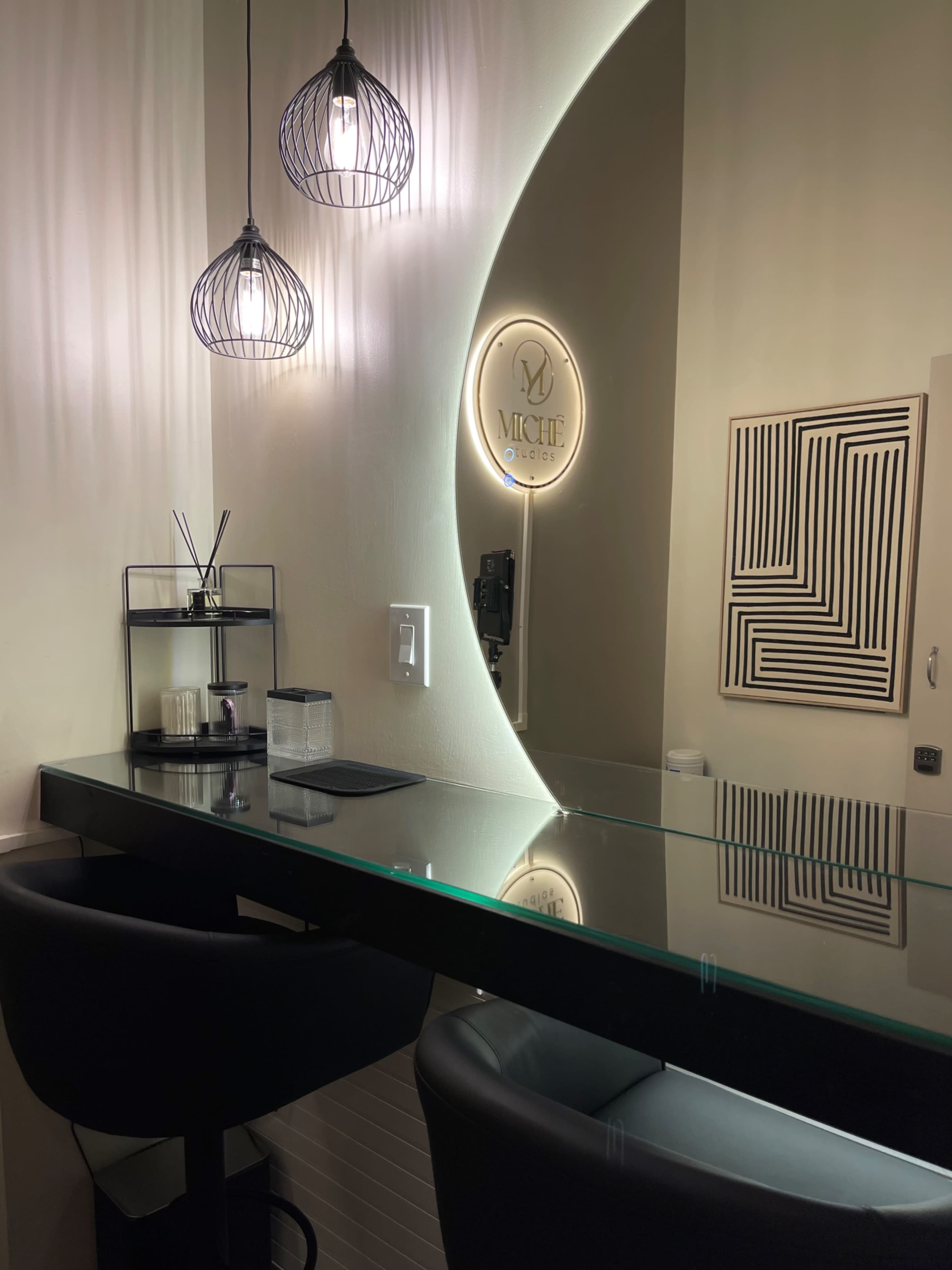 The image shows a minimalist beauty salon interior with a sleek glass table, two black chairs, and decorative elements including pendant lights and a wall-mounted sign.